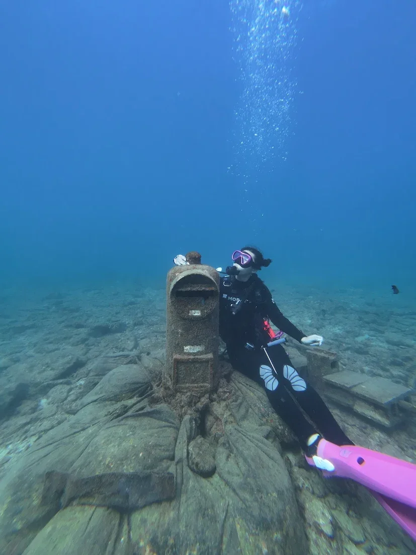 An underwater scene featuring a diver with a pink dive fin and purple mask posing next to a large, rusted mailbox on the ocean floor. The diver is holding a sketchbook and a pencil, and bubbles are rising from their breathing apparatus.