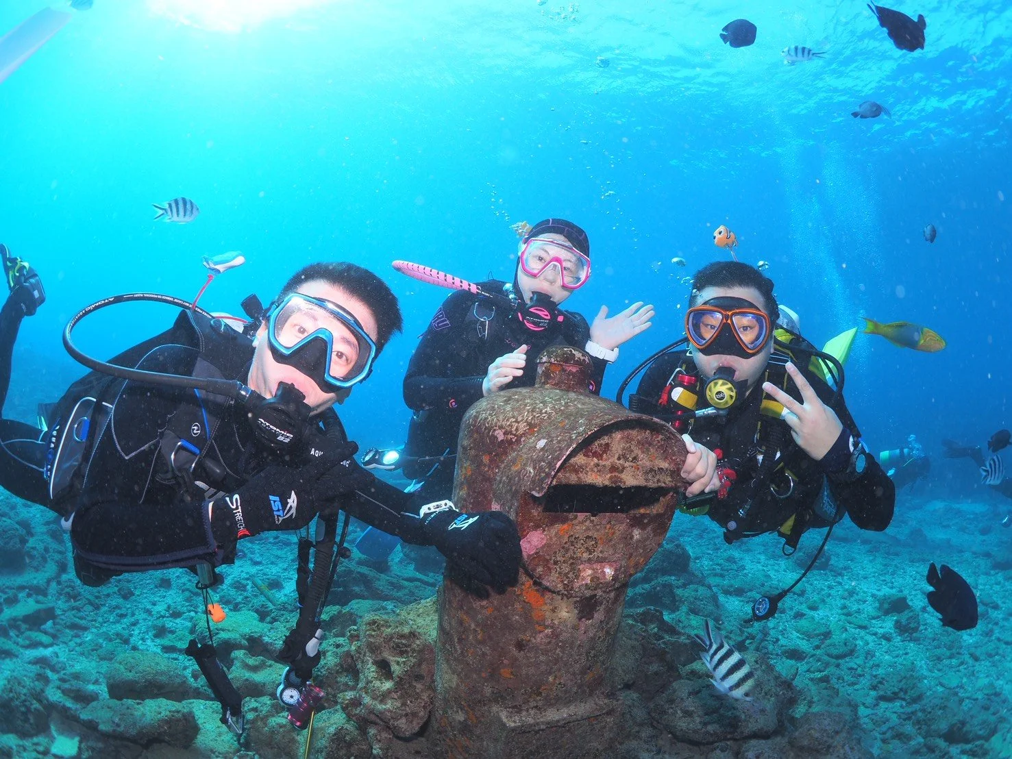 Three scuba divers underwater near a rusted pipe, surrounded by fish, making peace signs and looking at the camera.