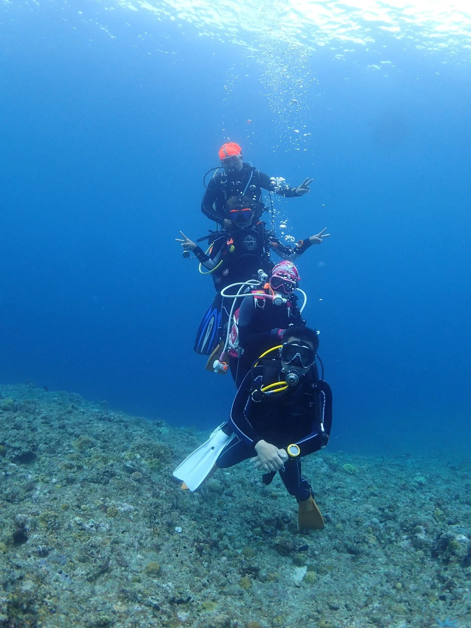 Two scuba divers pose underwater making peace signs while surrounded by coral and blue water.