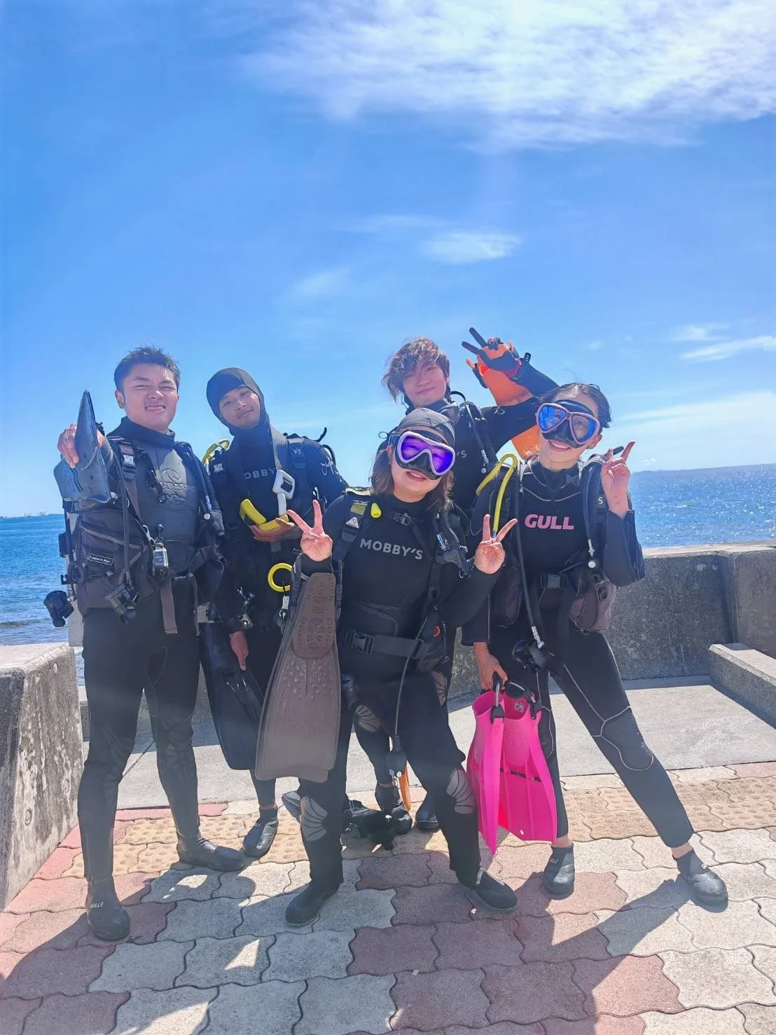 Group of five scuba divers in wetsuits and goggles, standing on a paved area by the ocean, smiling and making peace signs during daytime.