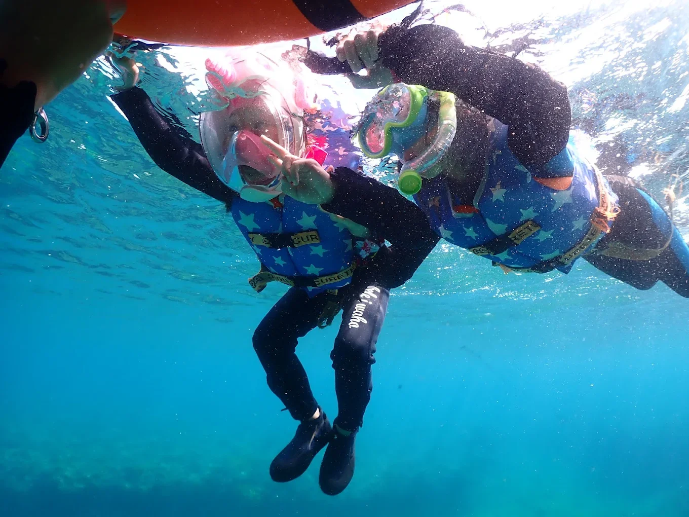 Two children in wetsuits and life jackets swimming underwater, with snorkeling masks, making peace signs.