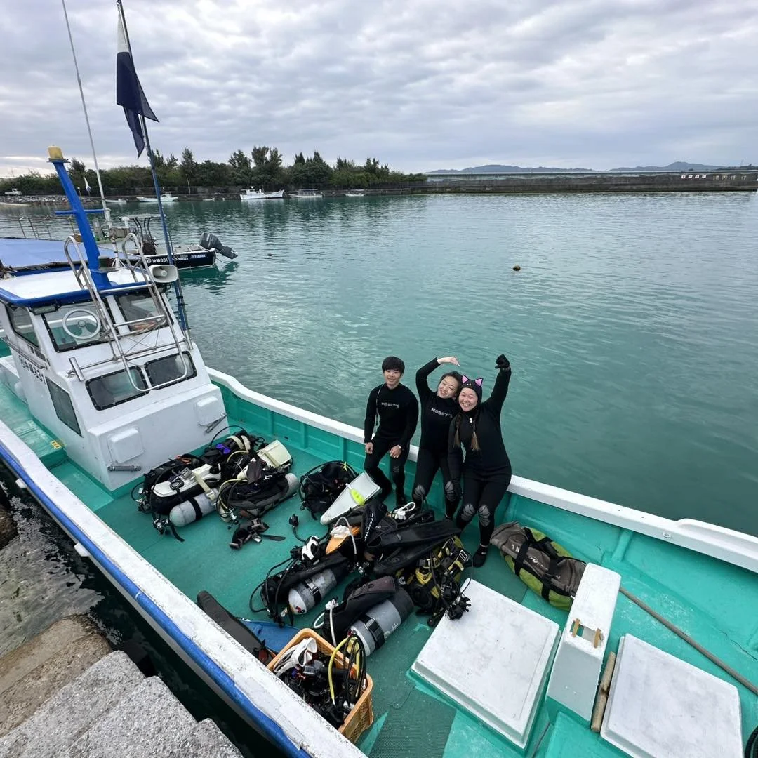 Three people in wetsuits standing on a boat with scuba diving gear, smiling and posing, near calm water and boats in the background.