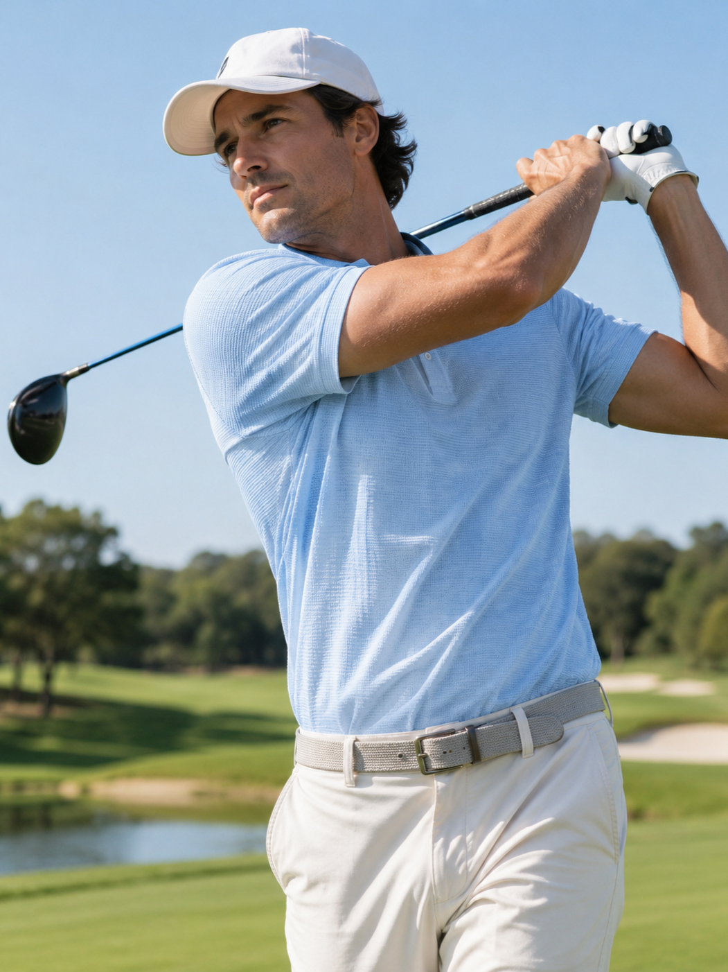 A man in a white cap, blue polo shirt, and beige pants swings a golf club on a golf course.