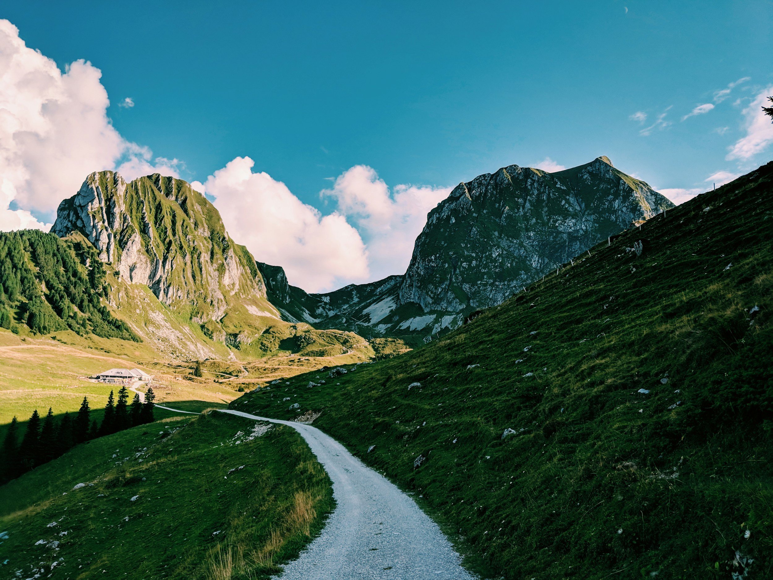 A mountainous landscape with a winding dirt path, lush green hills, and rugged mountain peaks under a bright blue sky with scattered clouds.