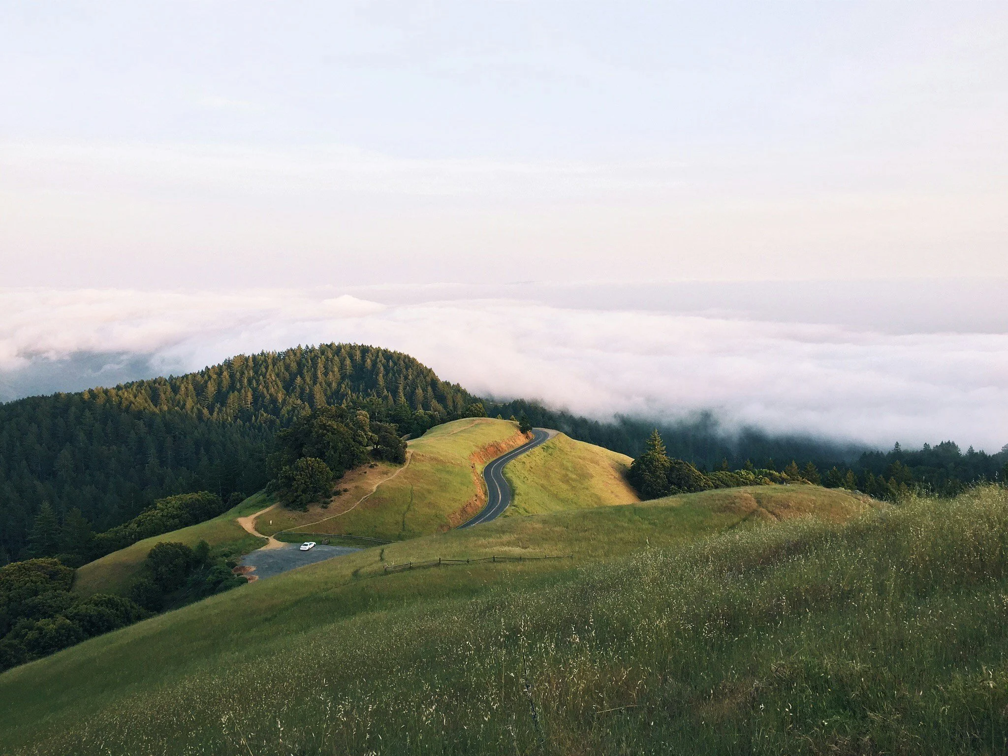 A scenic view of rolling green hills with a winding road and dense forest, under a sky filled with clouds.