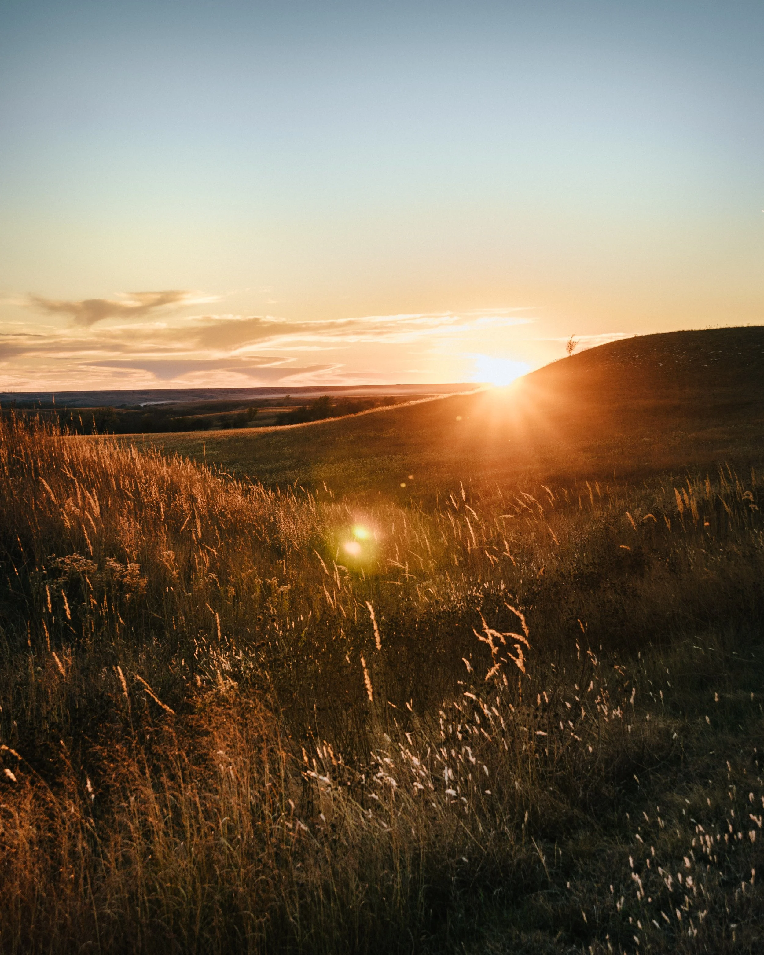 A landscape view of a sunset over grassy fields and rolling hills, with a partly cloudy sky.