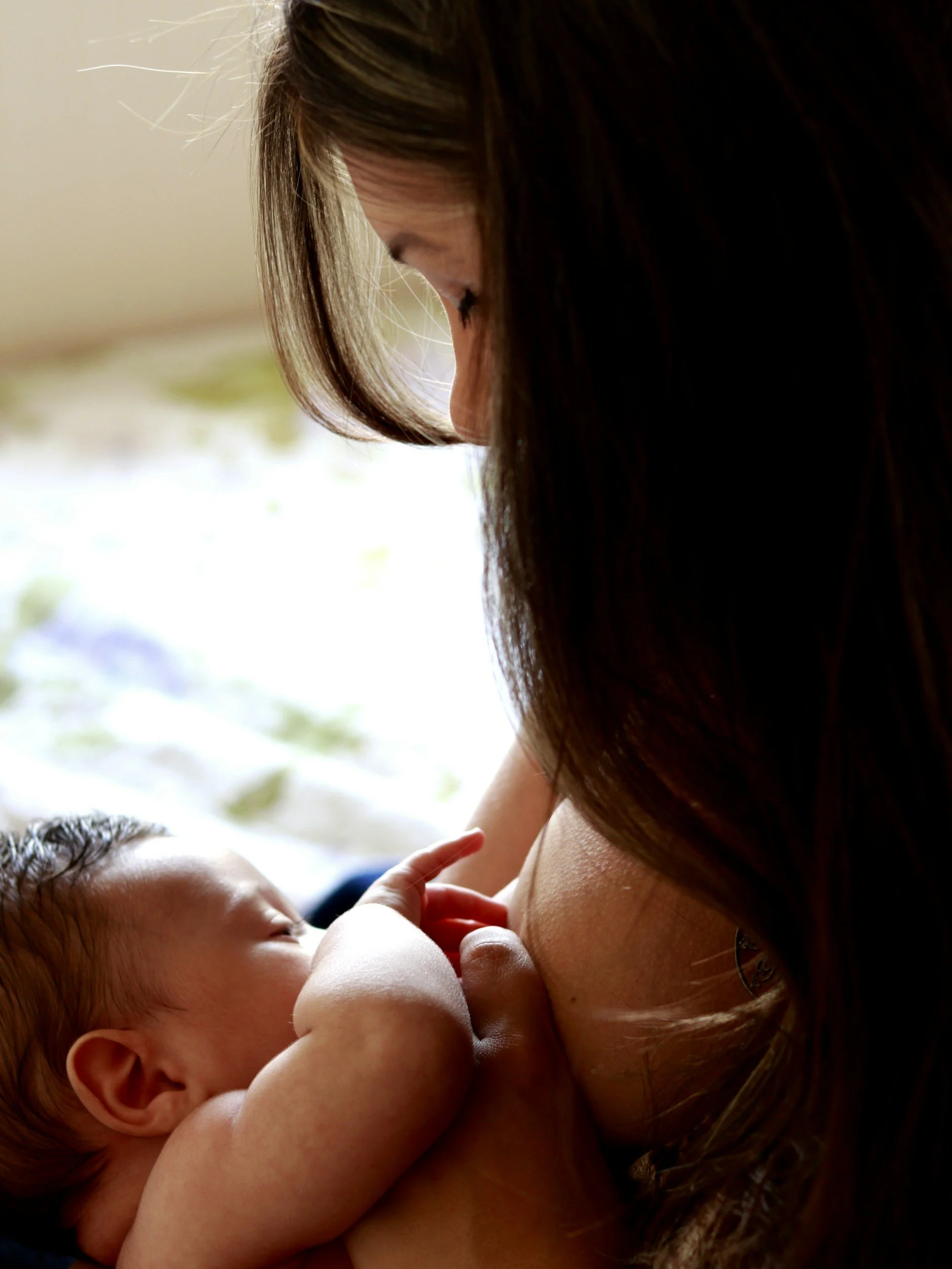 A woman with long brown hair is breastfeeding a baby, with the focus on their faces and the baby's hand touching the woman's chest.