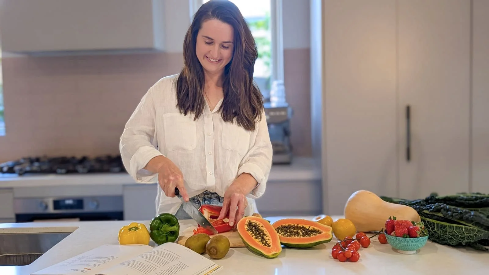 A woman in a white shirt slicing a red bell pepper at a kitchen counter, surrounded by colorful fruits and vegetables including papayas, strawberries, cherry tomatoes, and a butternut squash.