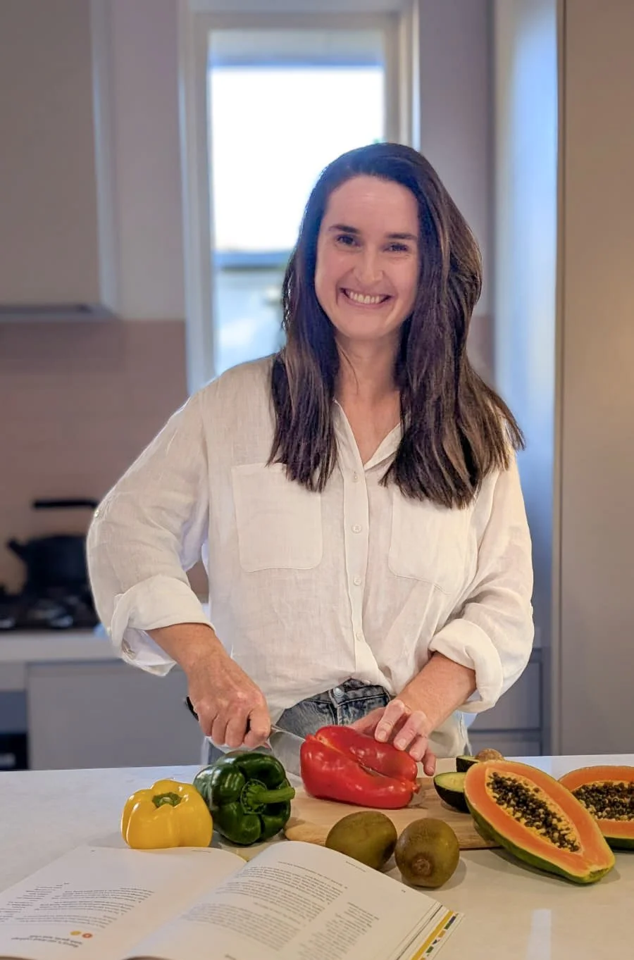 A woman with long brown hair, smiling and chopping a red bell pepper in a modern kitchen. On the counter are a yellow bell pepper, green bell pepper, two kiwis, and two halved papayas, with an open cookbook nearby.