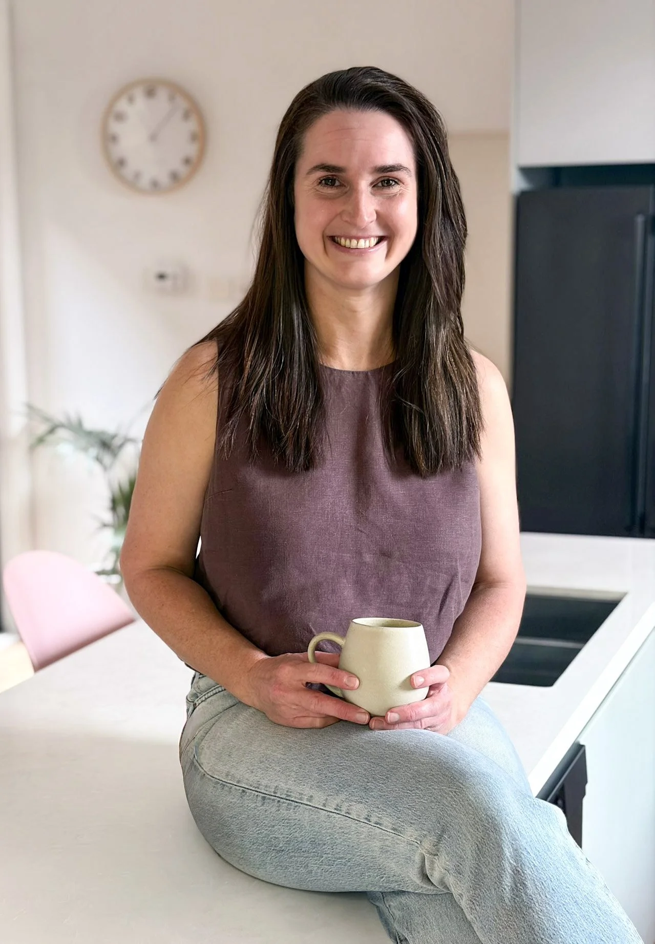 A woman with long dark hair smiling, sitting cross-legged on a white kitchen counter, holding a beige mug. She is wearing a sleeveless maroon top and light gray jeans. In the background, there is a wall clock showing approximately 1:02 and a blurred plant.