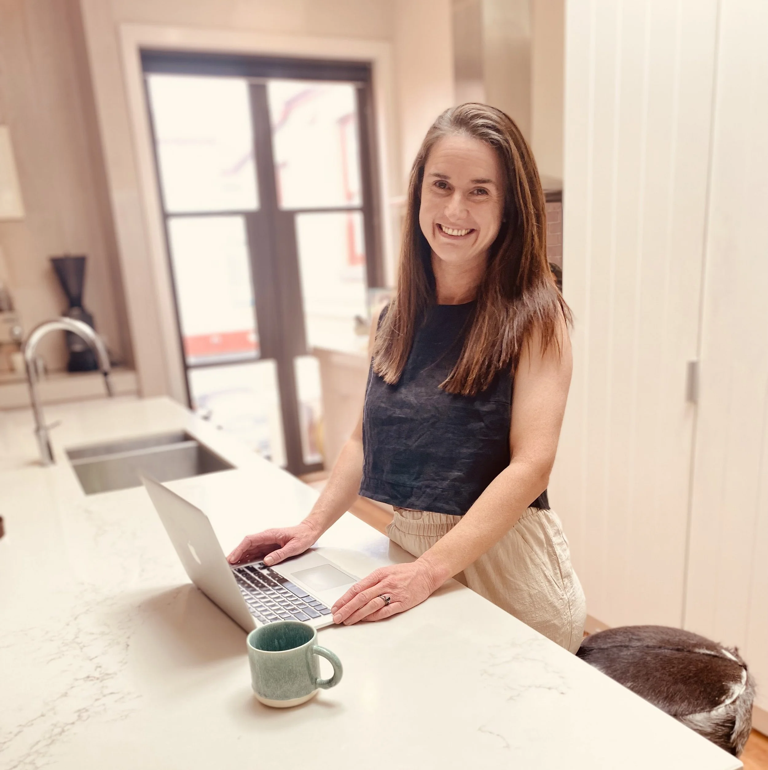 Woman standing at kitchen counter using laptop, green mug on counter, dog lying on the floor nearby.