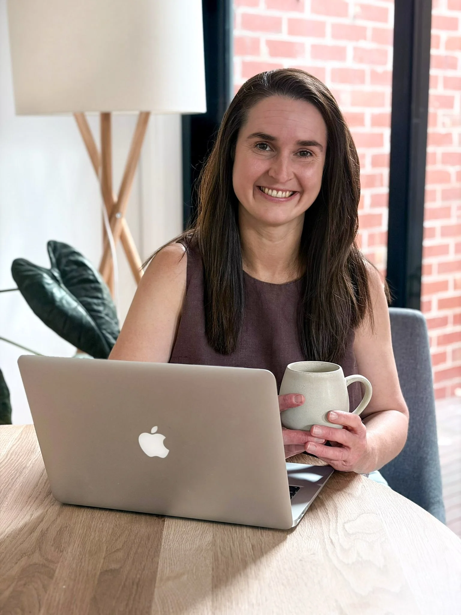 A woman with long brown hair smiling while sitting at a wooden table with a silver MacBook laptop and holding a coffee mug. Behind her is a glass door with a brick wall outside and a large beige floor lamp.