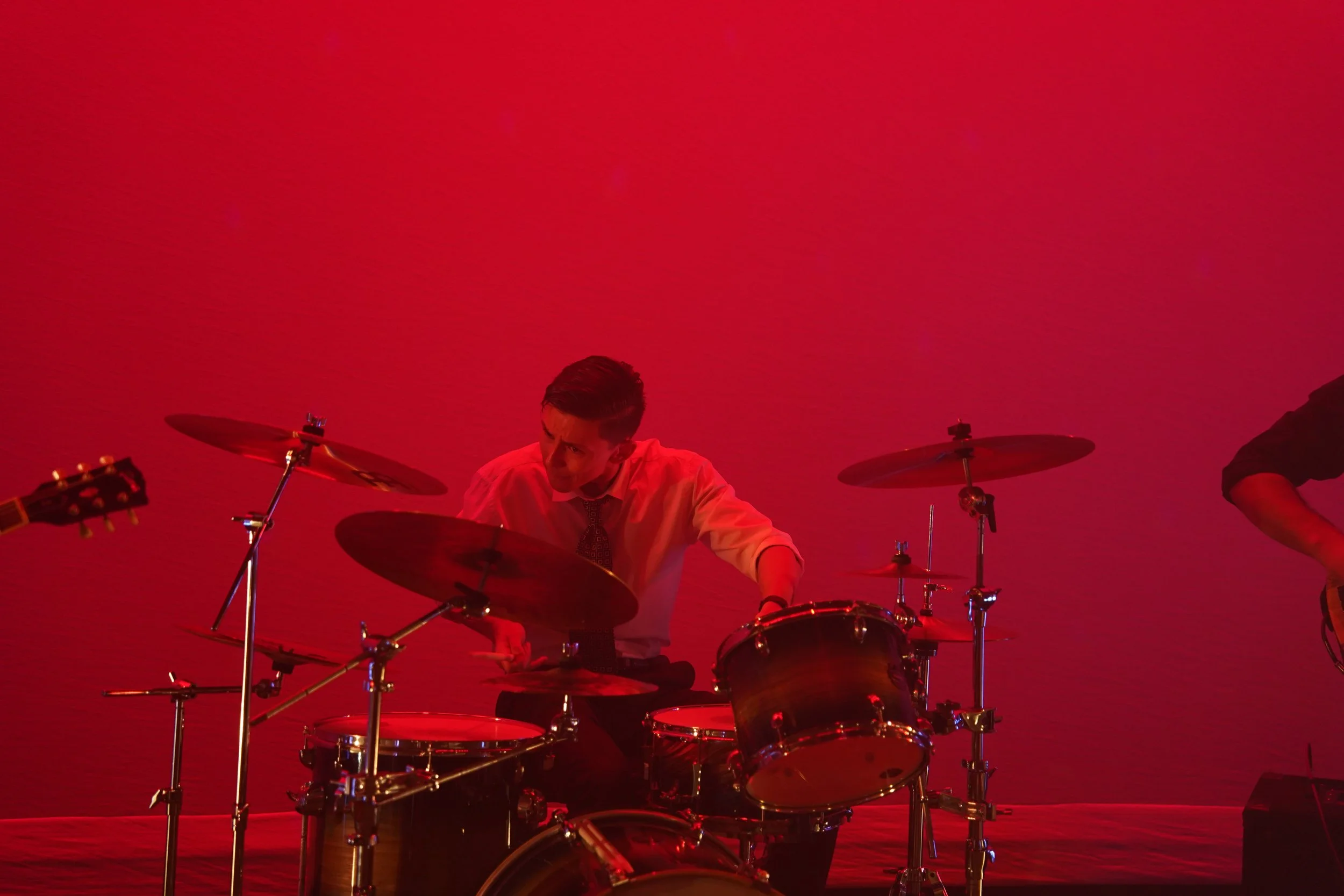 A person playing a drum set on stage with a red background.