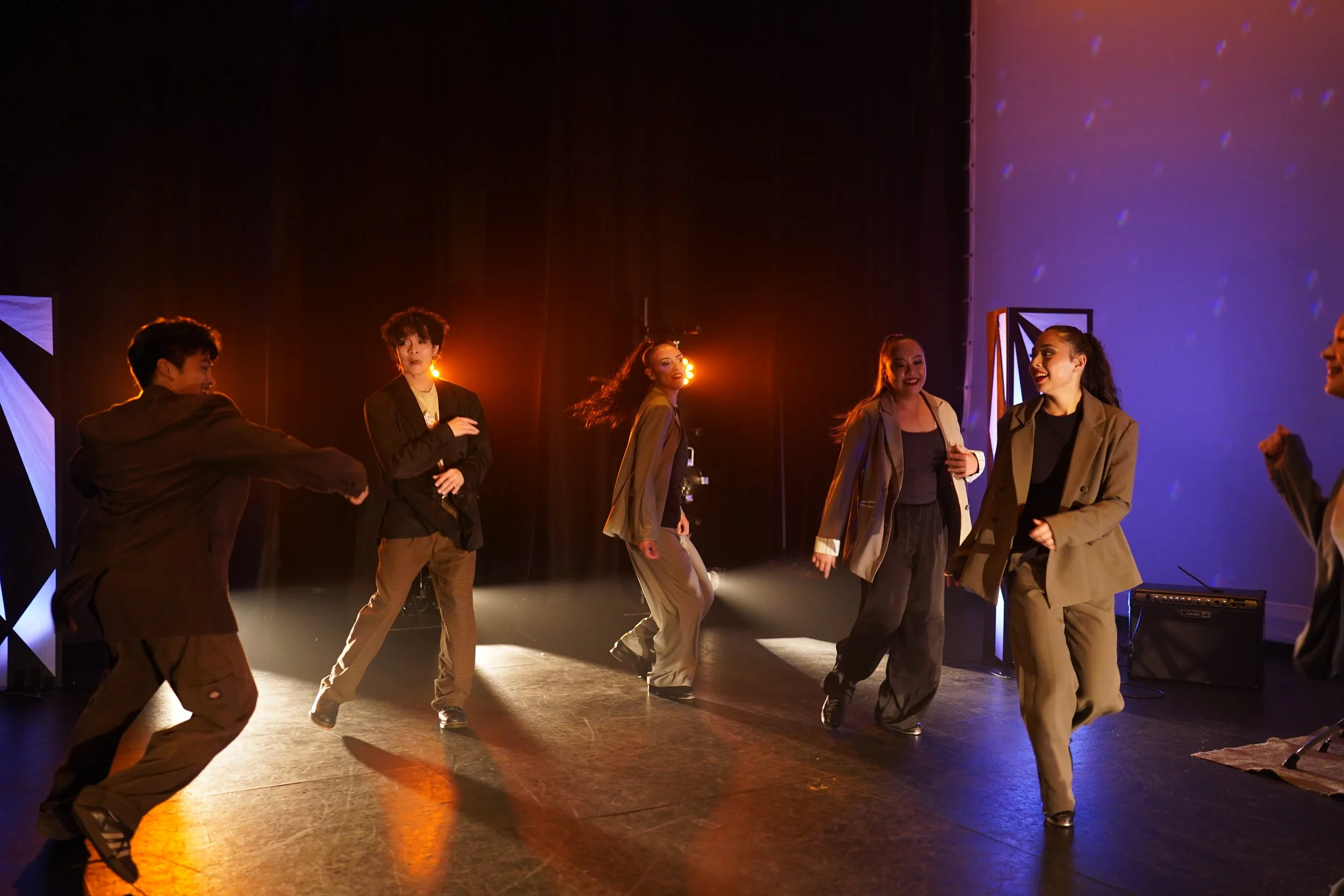 Group of six diverse dancers performing on stage with warm lighting and colorful backdrop.