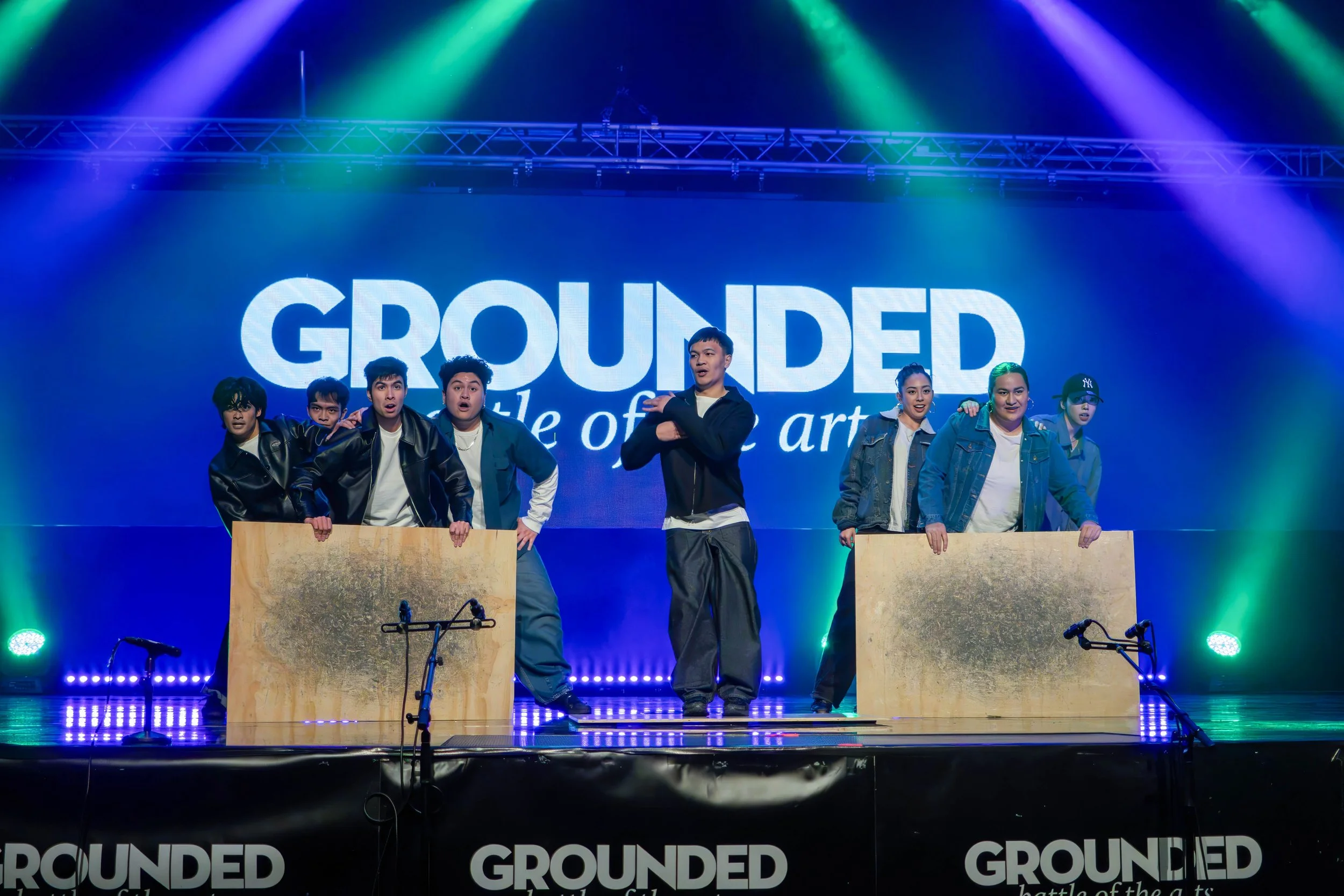 A group of six people on stage participating in a performance or event, with a large screen behind them displaying the word 'GROUNDED' and the phrase 'battle of the arts', under colorful stage lighting.