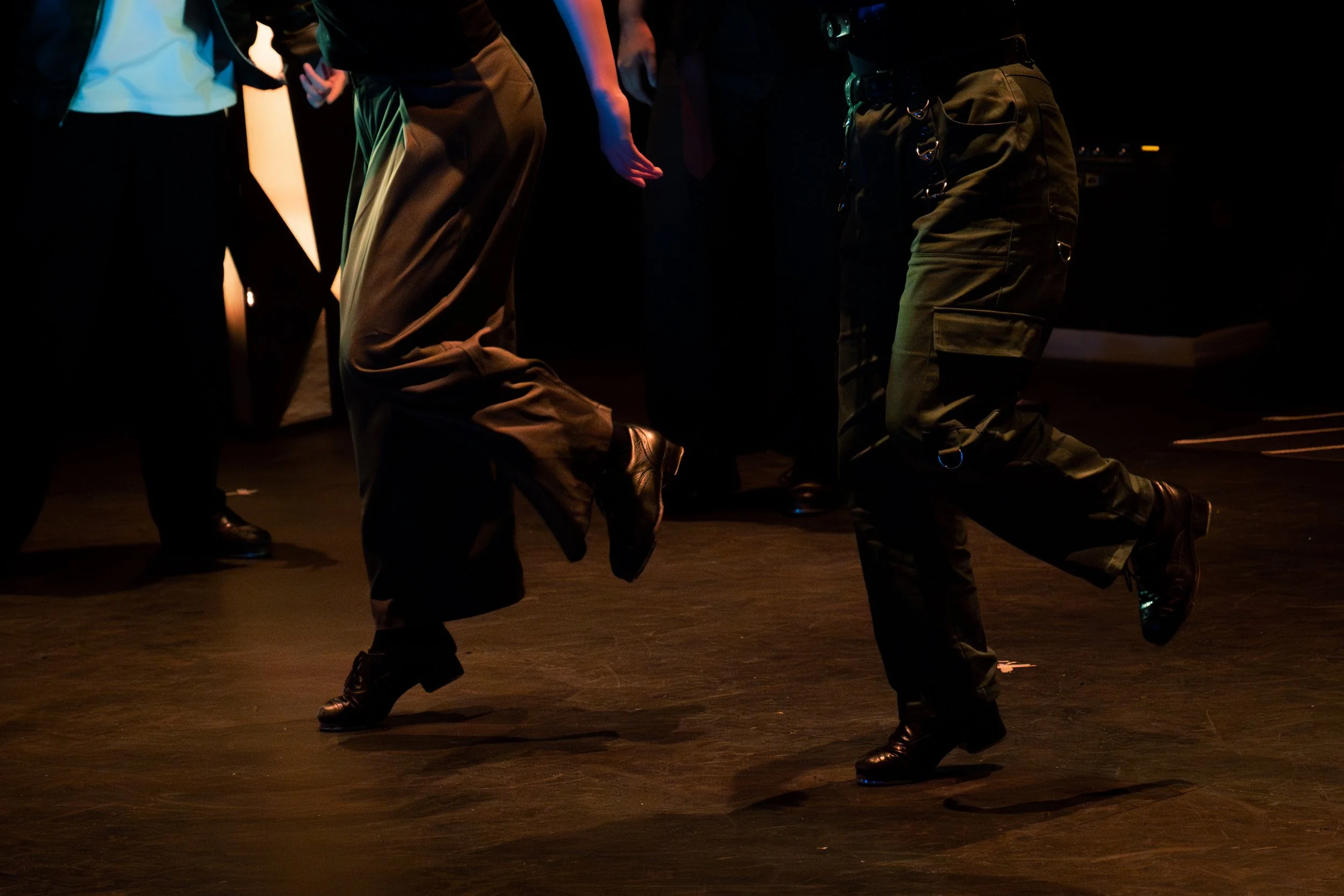 People performing a dance with bent knees, wearing dark clothing and boots, on a dimly lit stage.