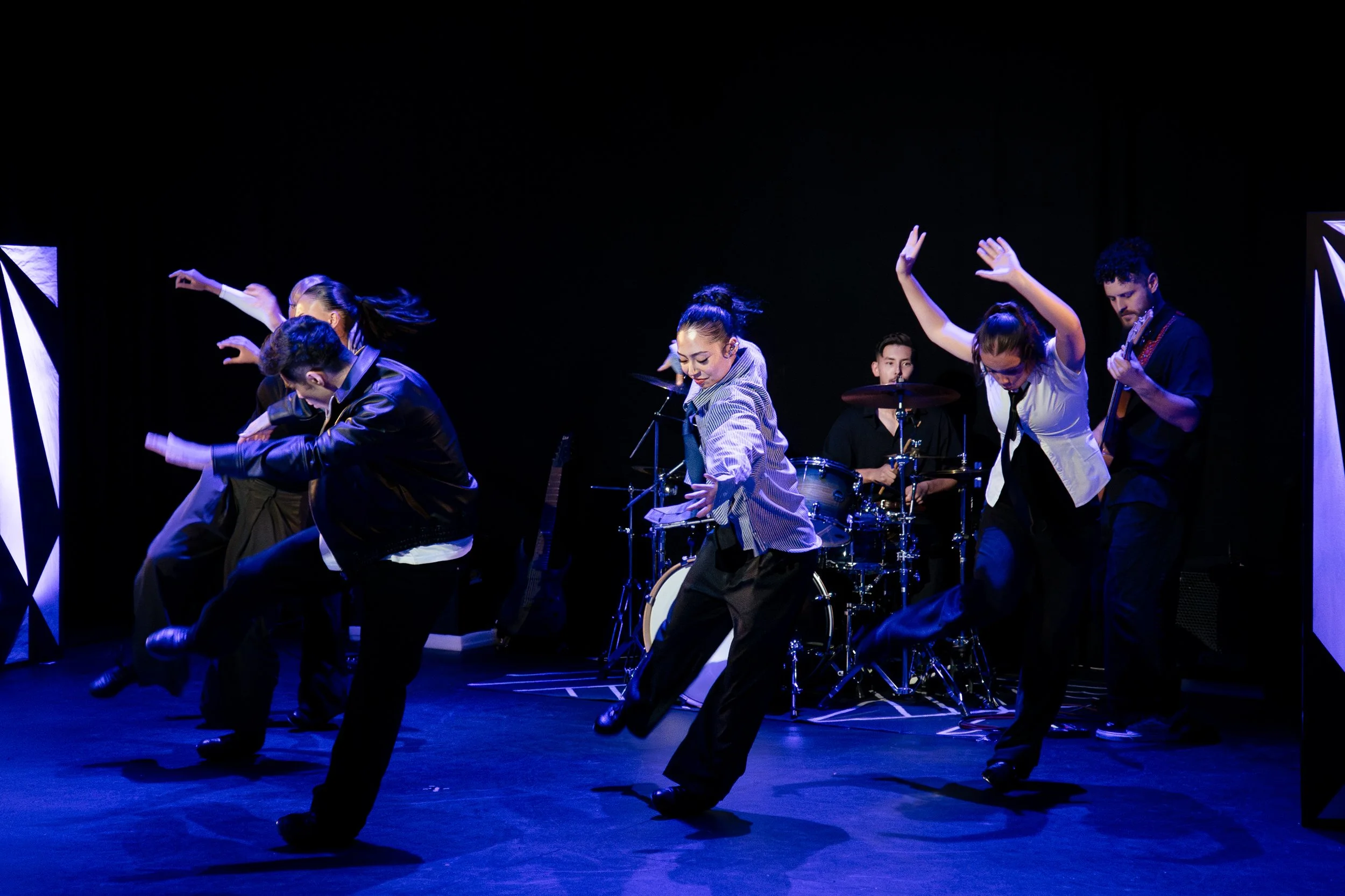 Group of performers dancing on stage with a band playing in the background, illuminated by blue stage lighting.