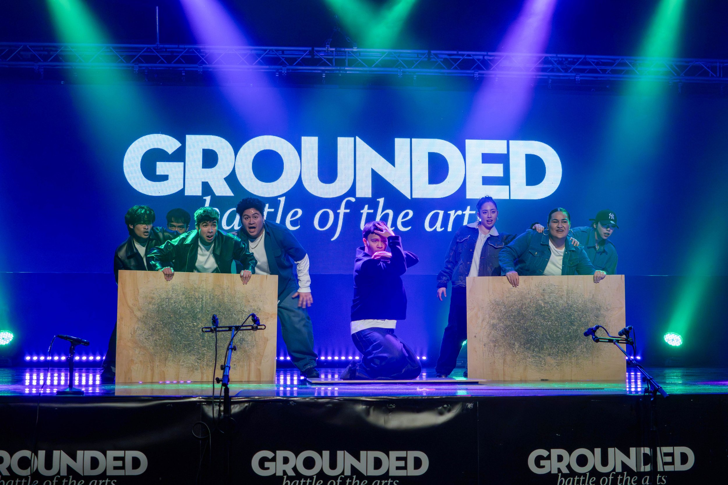 Performance on stage with seven performers behind wooden panels, colorful stage lights, and a large screen displaying the word 'GROUNDED' and the phrase 'battle of the arts'.