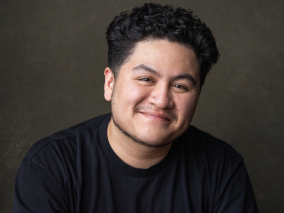 A young man with dark curly hair smiling at the camera, wearing a black t-shirt, against a plain dark background.