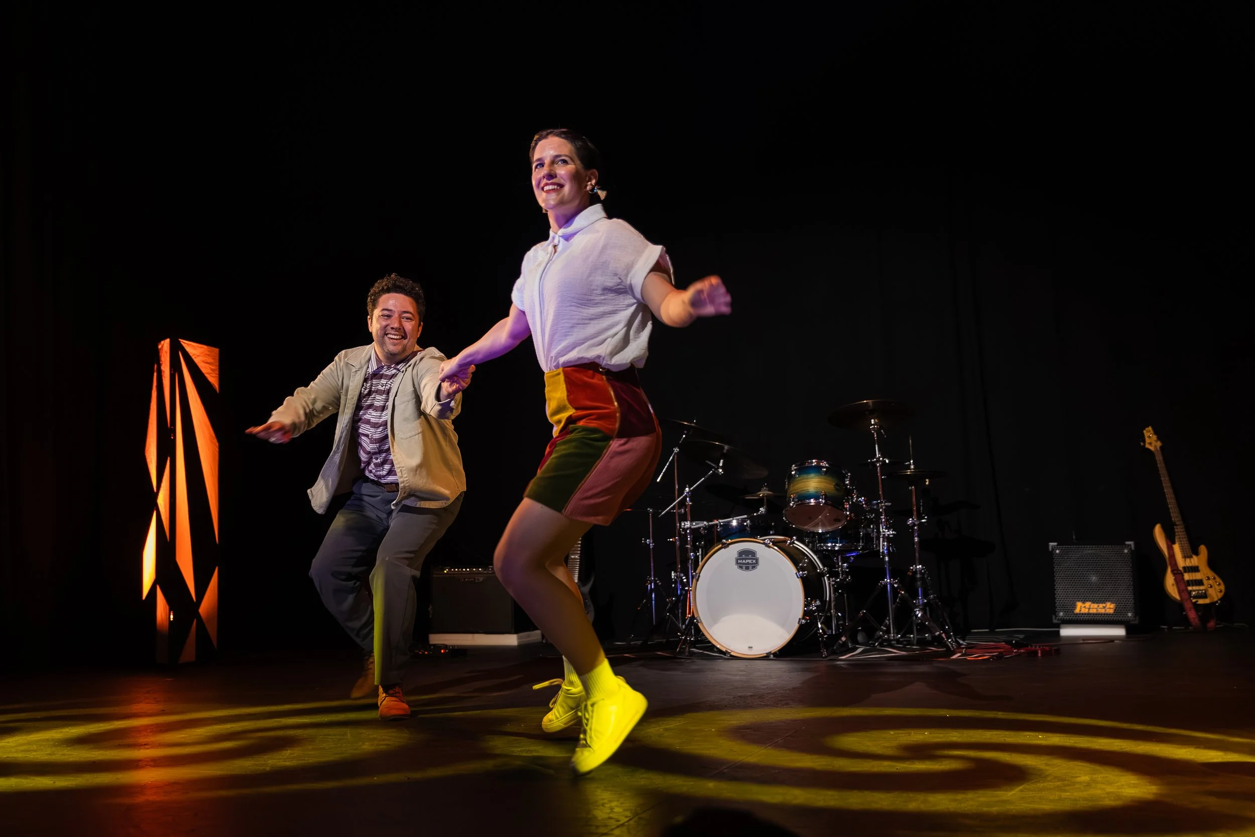 Two performers dancing on a stage with musical instruments in the background, illuminated by stage lights.