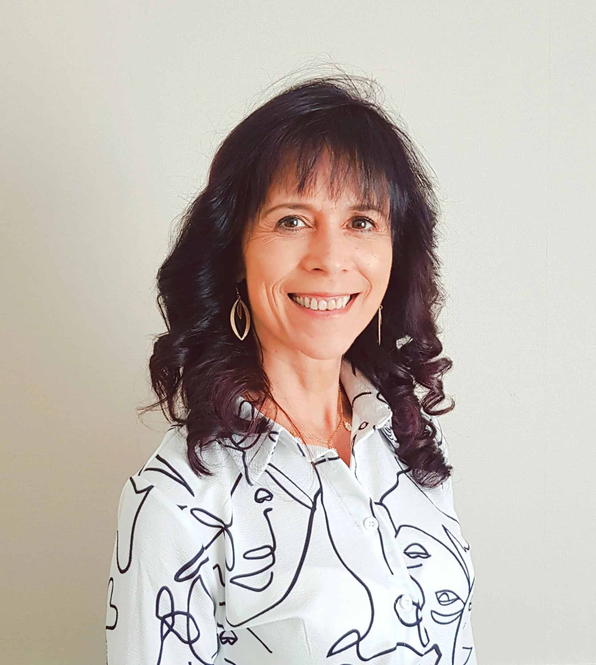 A smiling woman with dark curly hair wearing earrings and a patterned blouse stands against a plain light-colored wall.