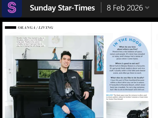 A young man with black hair, wearing a black jacket and blue jeans, sitting on a bench next to a black piano in a modern room, smiling at the camera.