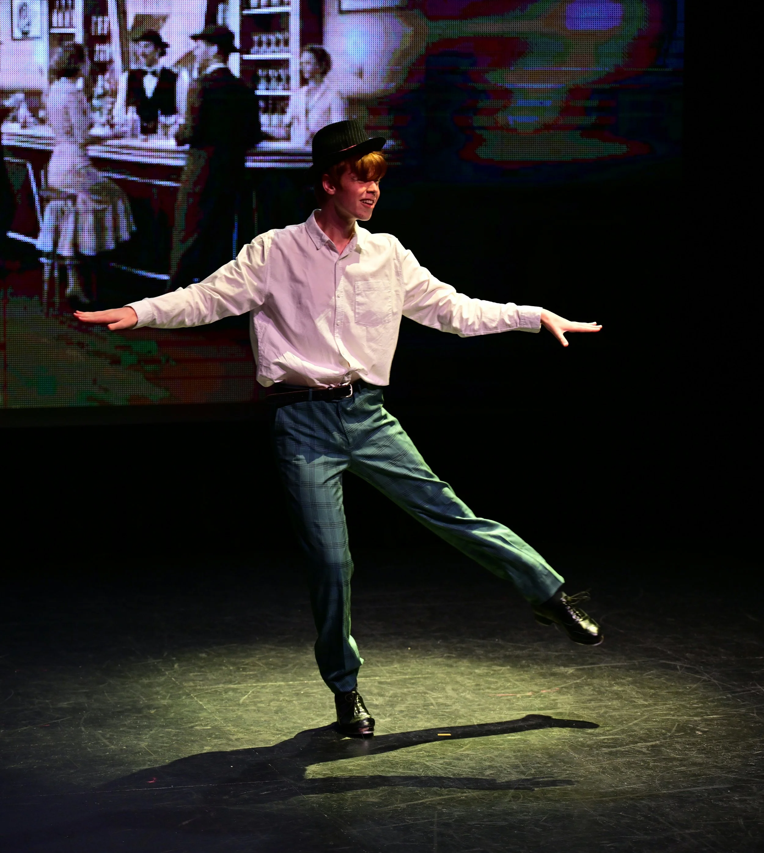 Performer in a white shirt, plaid pants, black hat, and black shoes dancing on a stage with a colorful digital backdrop.