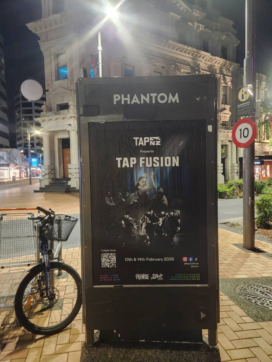 Nighttime street scene with a poster advertising 'Tap Fusion' event, happening on February 13th and 14th, 2026. The poster features dancers performing on stage. A bicycle is parked next to the poster and nearby are buildings, streetlights, and a no p