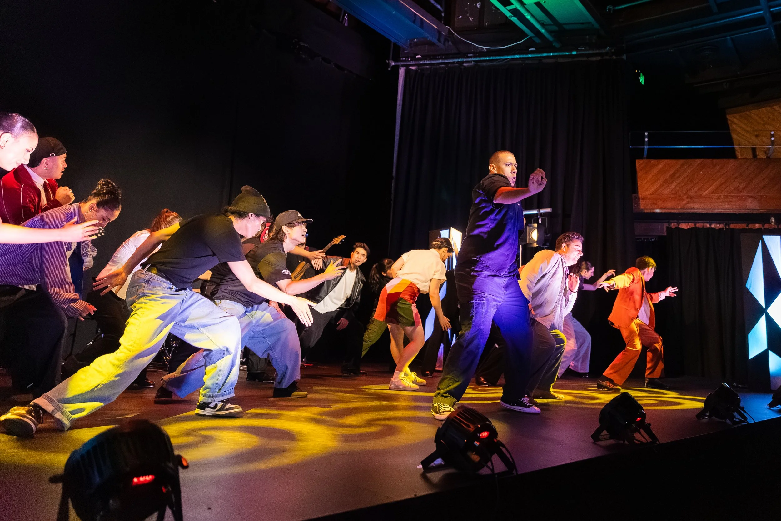 Group of performers on stage dancing, dressed in colorful clothing, with lighting and stage equipment visible.