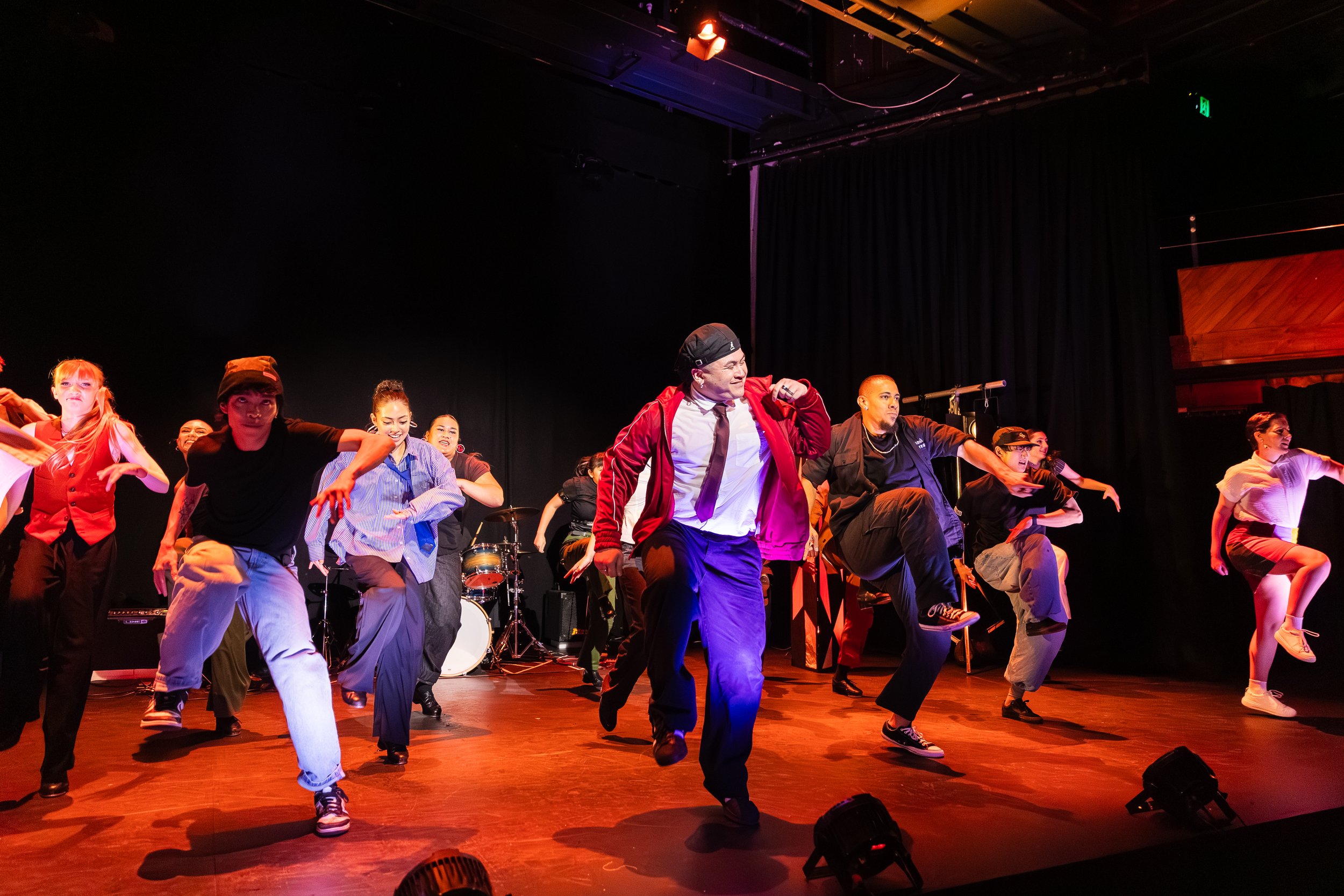 A diverse group of dancers performing on stage with a black backdrop and musical instruments in the background.