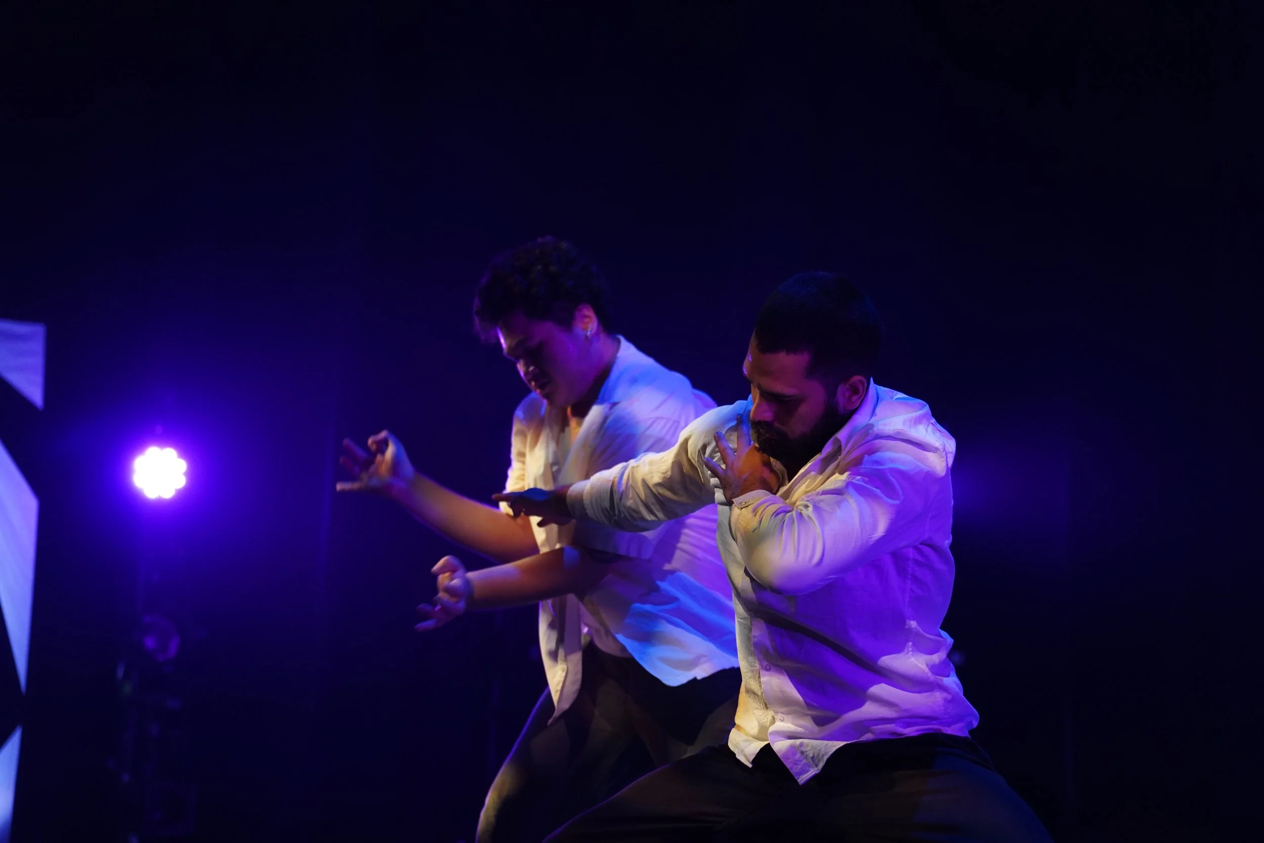 Two men wearing white shirts are on stage, knees bent, with expressive facial expressions, illuminated by purple stage lighting.