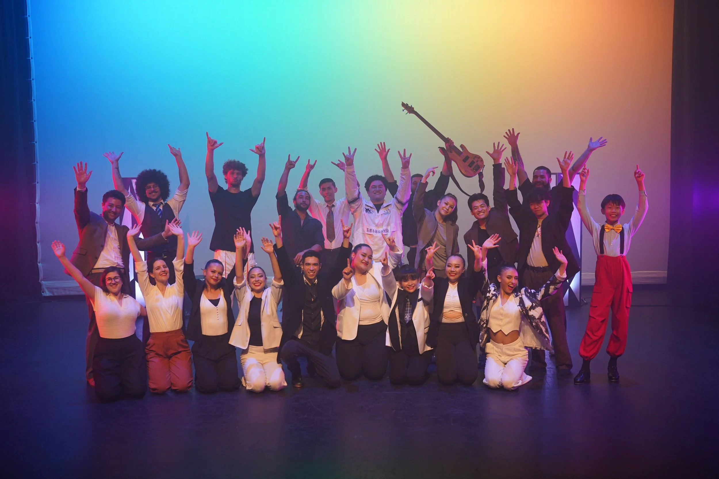Group of people on stage celebrating, some making victory signs, with a rainbow-colored background. A guitarist is being lifted, and everyone is smiling and posing happily.