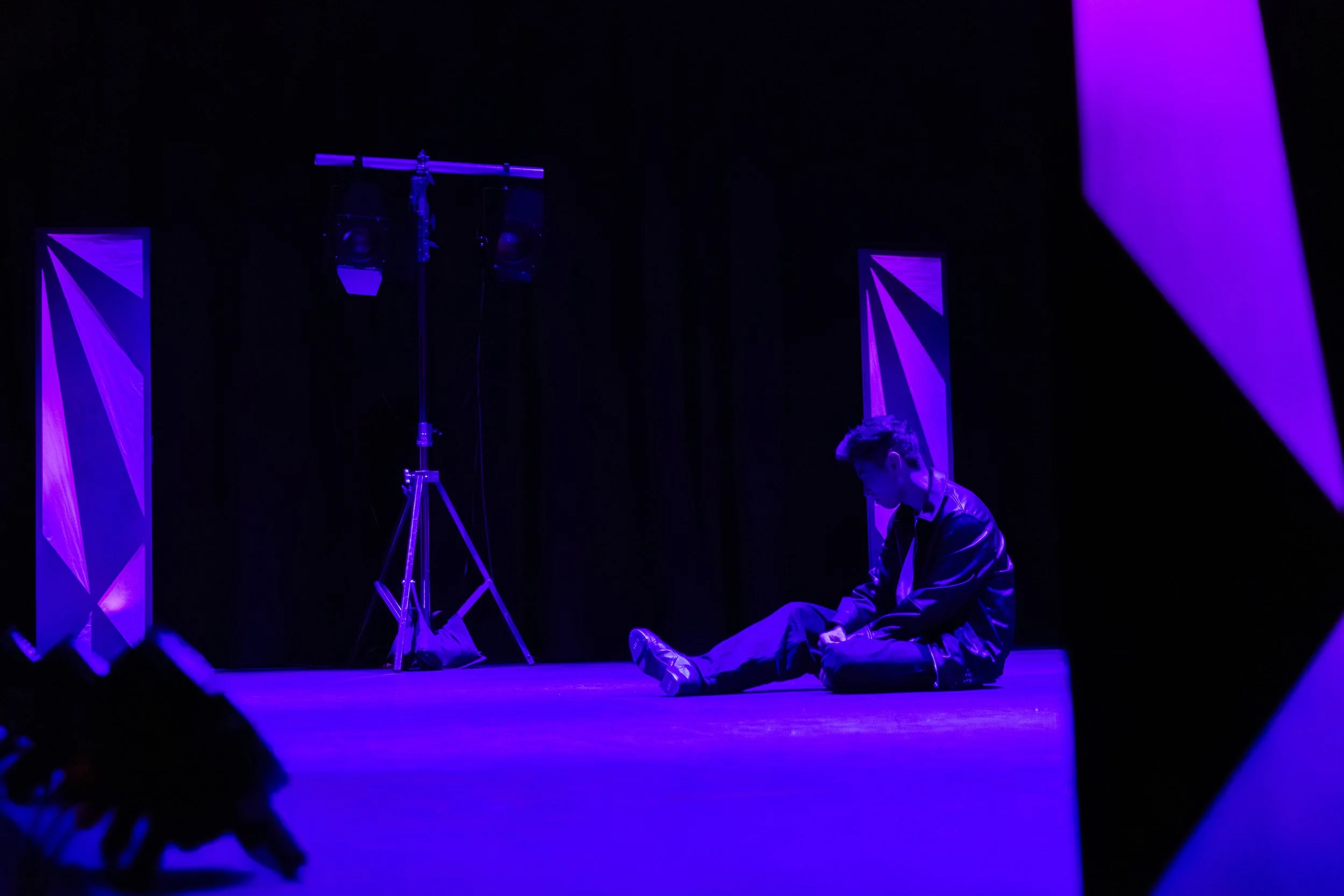 A person sitting on stage in dim purple lighting, with a microphone stand and stage decor in the background.