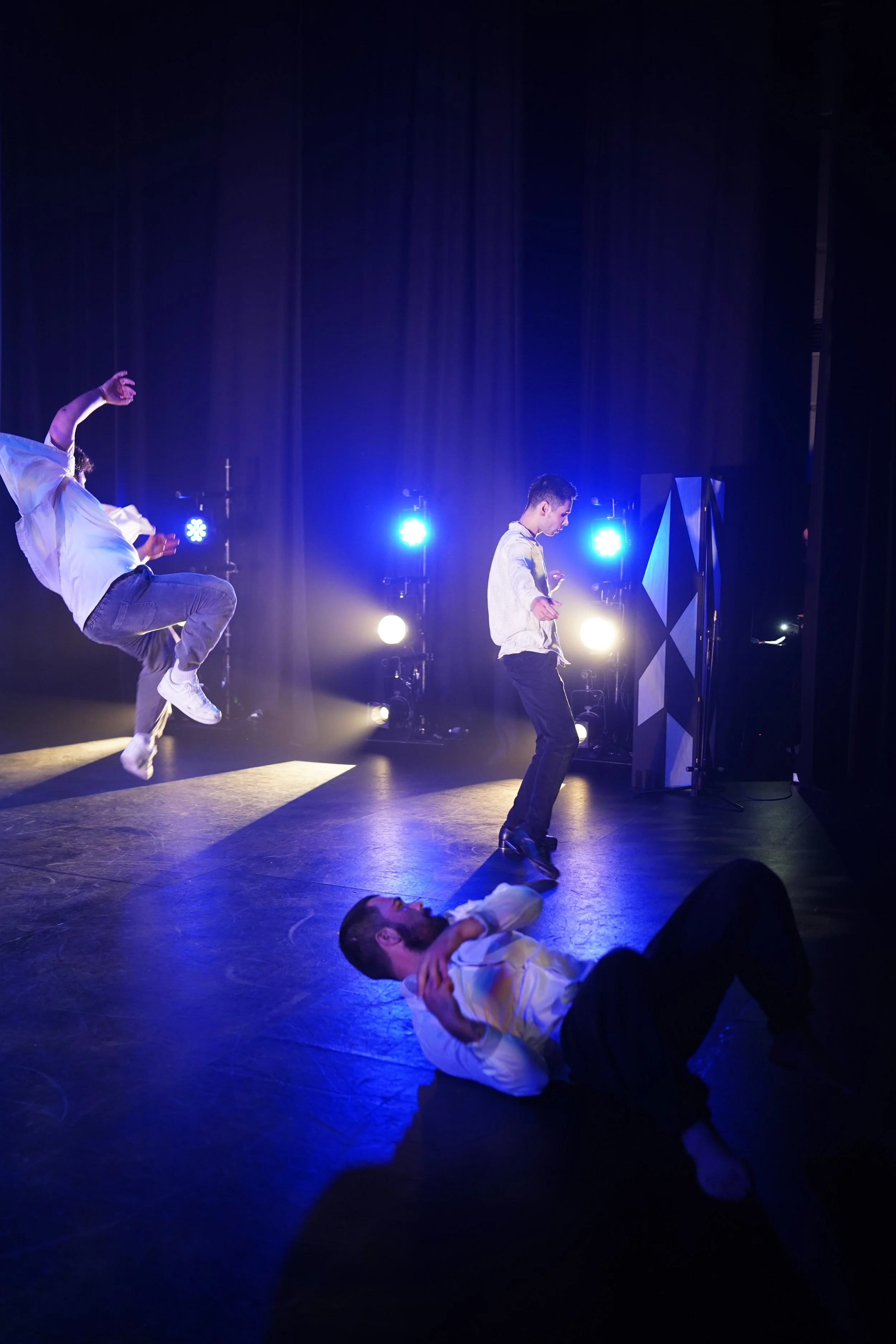 Four performers on stage under blue and white stage lights, one lying on the floor in a white shirt with hands over ears, another mid-air jump with a white shirt and gray pants, a third standing with a microphone, and the fourth partially visible beh
