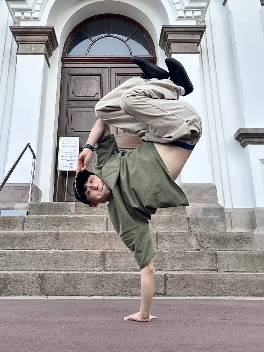 A young man performing a handstand with one hand on the ground, balancing his body with his legs bent and crossed at the ankles, on the steps outside a building with white walls and a wooden door.