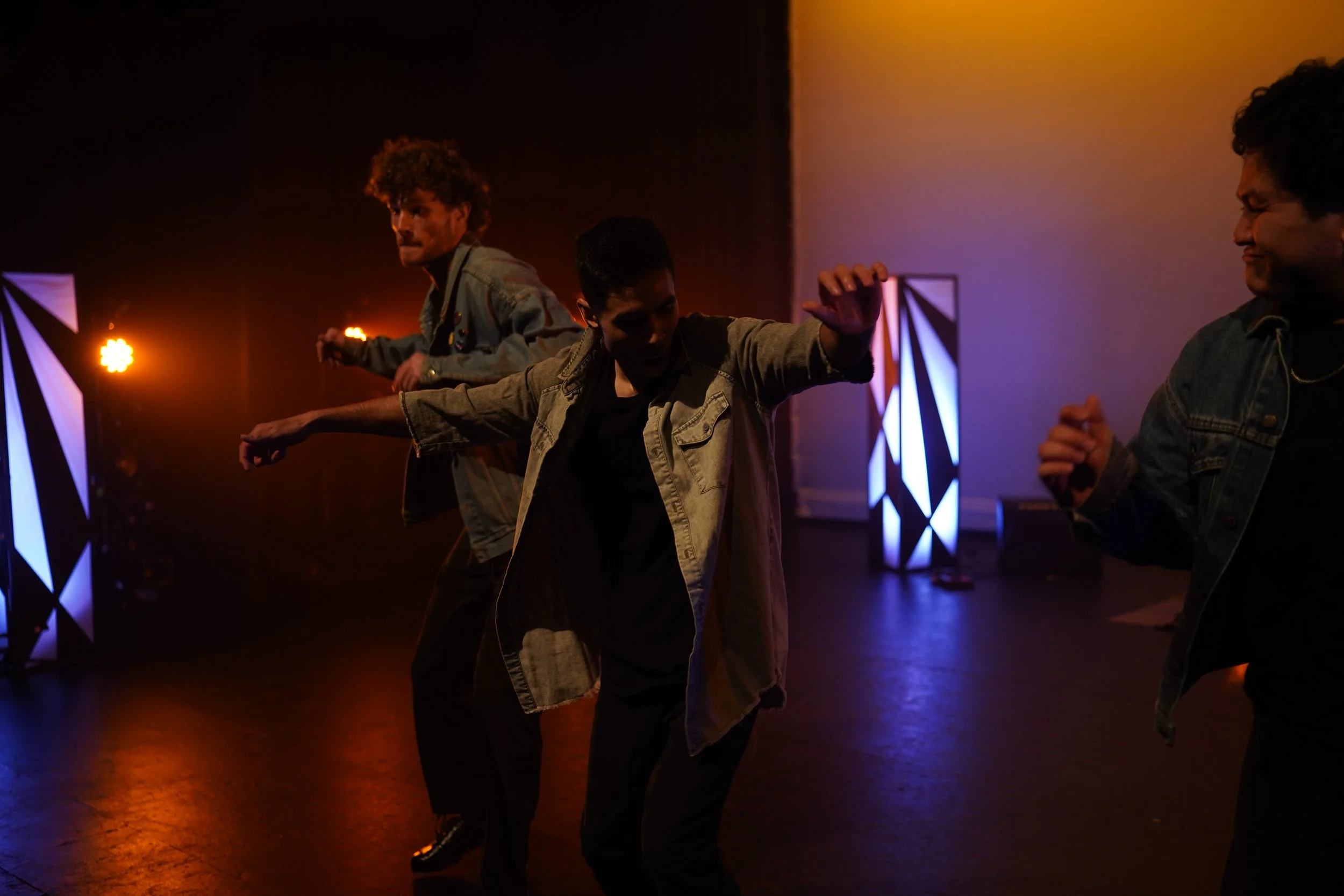 Four young people dancing together in a dimly lit indoor setting with colorful stage lights and geometric light fixtures in the background.