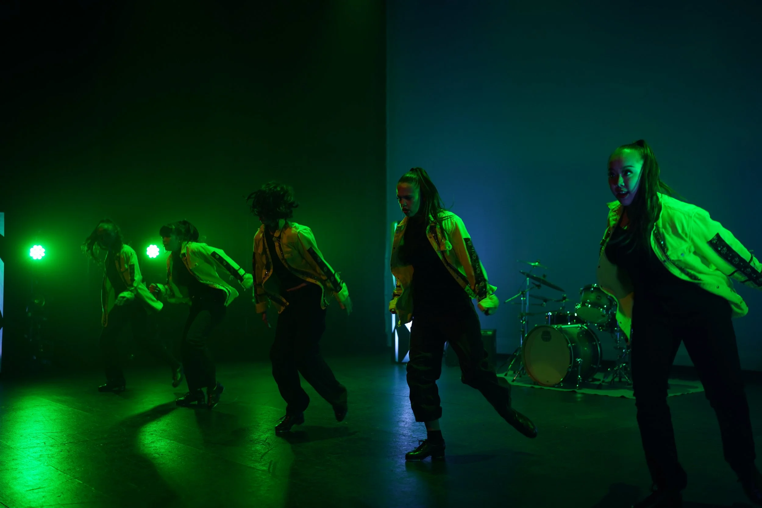 A group of performers dressed in yellow jackets and dark pants dancing on stage under green lighting, with a drum set visible in the background.