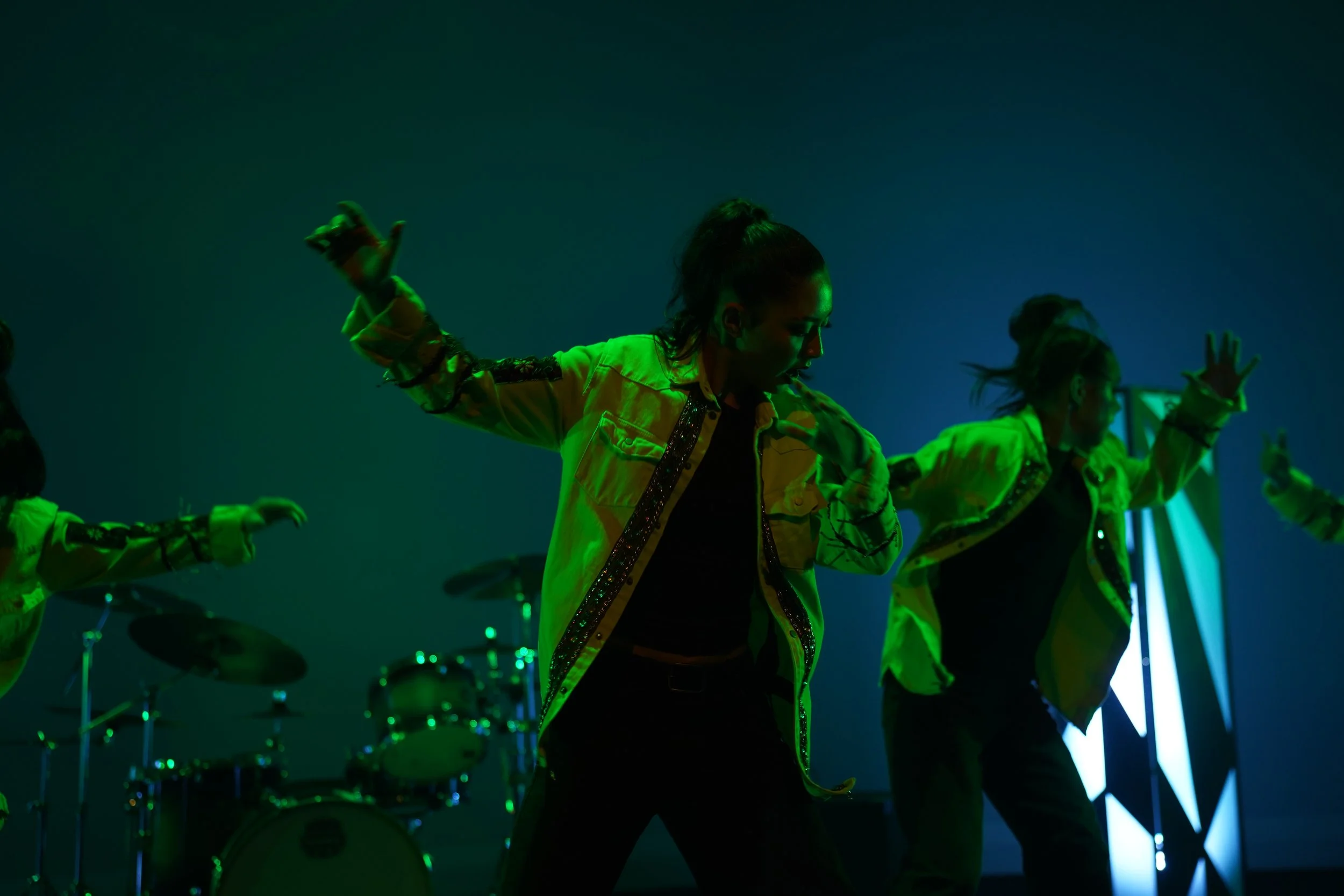 Dancers in neon yellow jackets performing under green and blue stage lighting, with drums in the background.