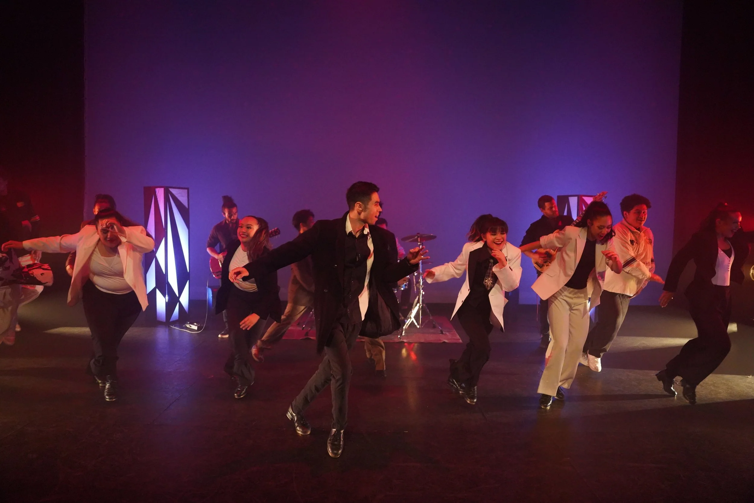 A group of performers dancing on stage with colorful lighting, some playing musical instruments in the background.