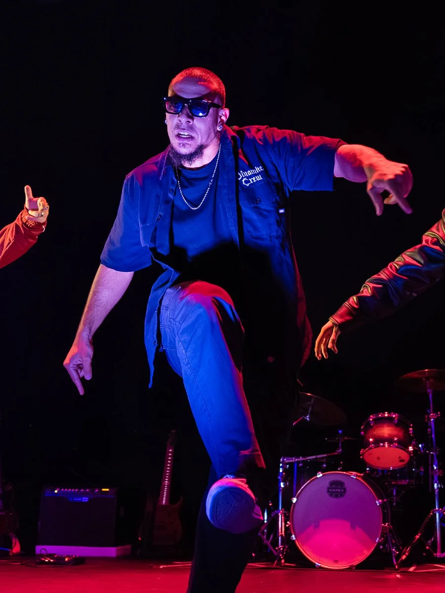 Male dancer performing on stage in a black shirt with sunglasses, cutting a dynamic pose with one knee up, illuminated by colorful stage lights.