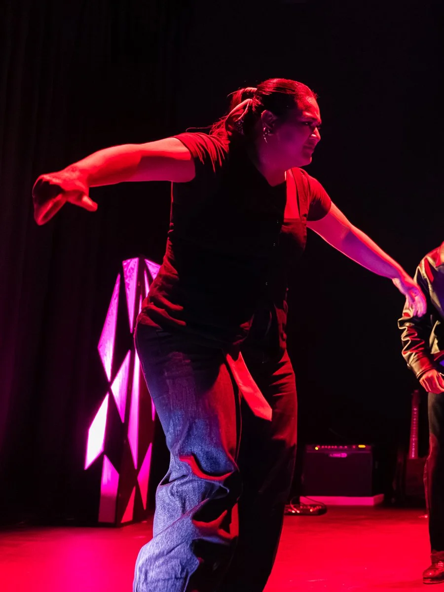 A woman with dark hair tied back, wearing a black shirt and jeans, is performing on stage under red and pink stage lighting. She is leaning forward with her arms extended, appearing to be in the middle of a dance or theatrical performance.