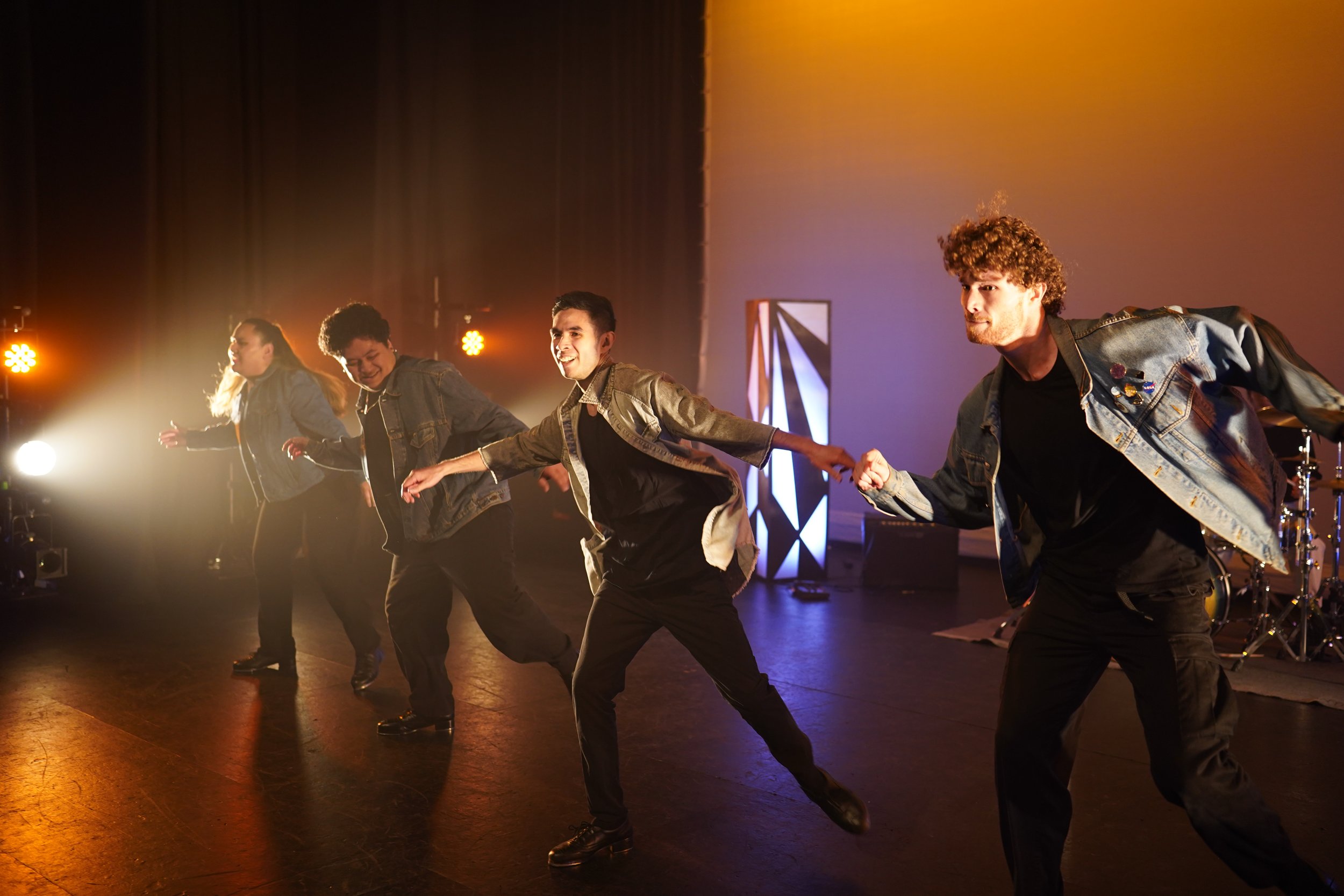 A group of five young adults dancing together on a stage with warm lighting and musical instruments in the background.