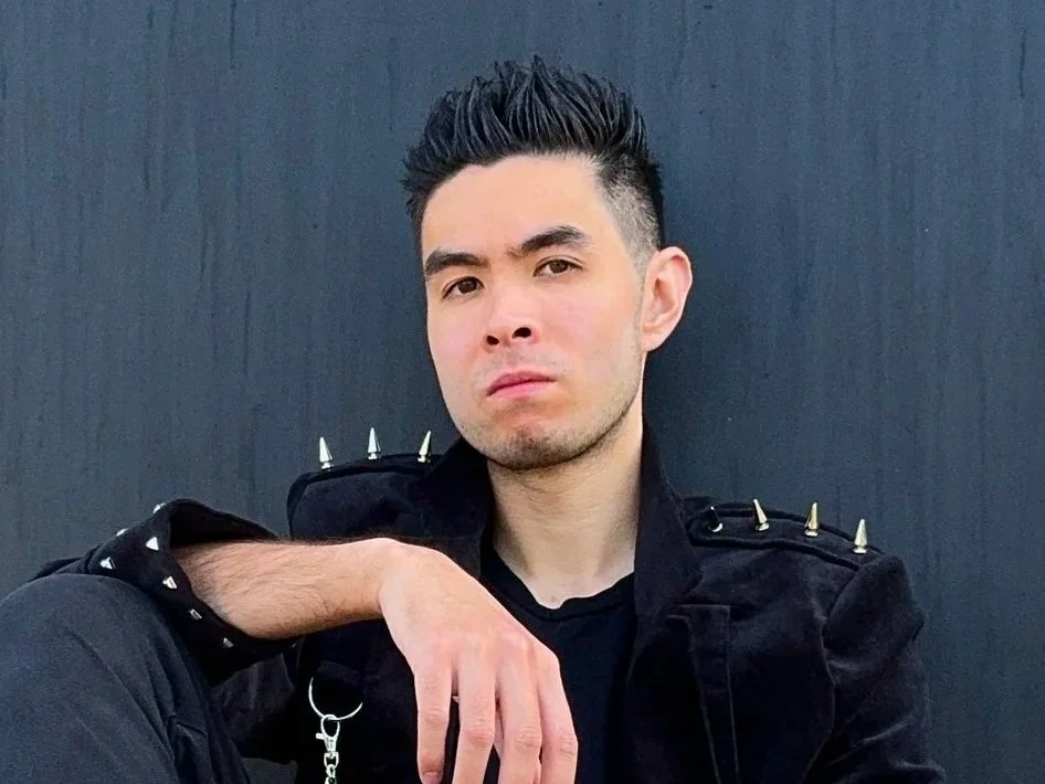 A young man with styled black hair, sitting against a dark background, wearing a black jacket with spikes on the shoulders.