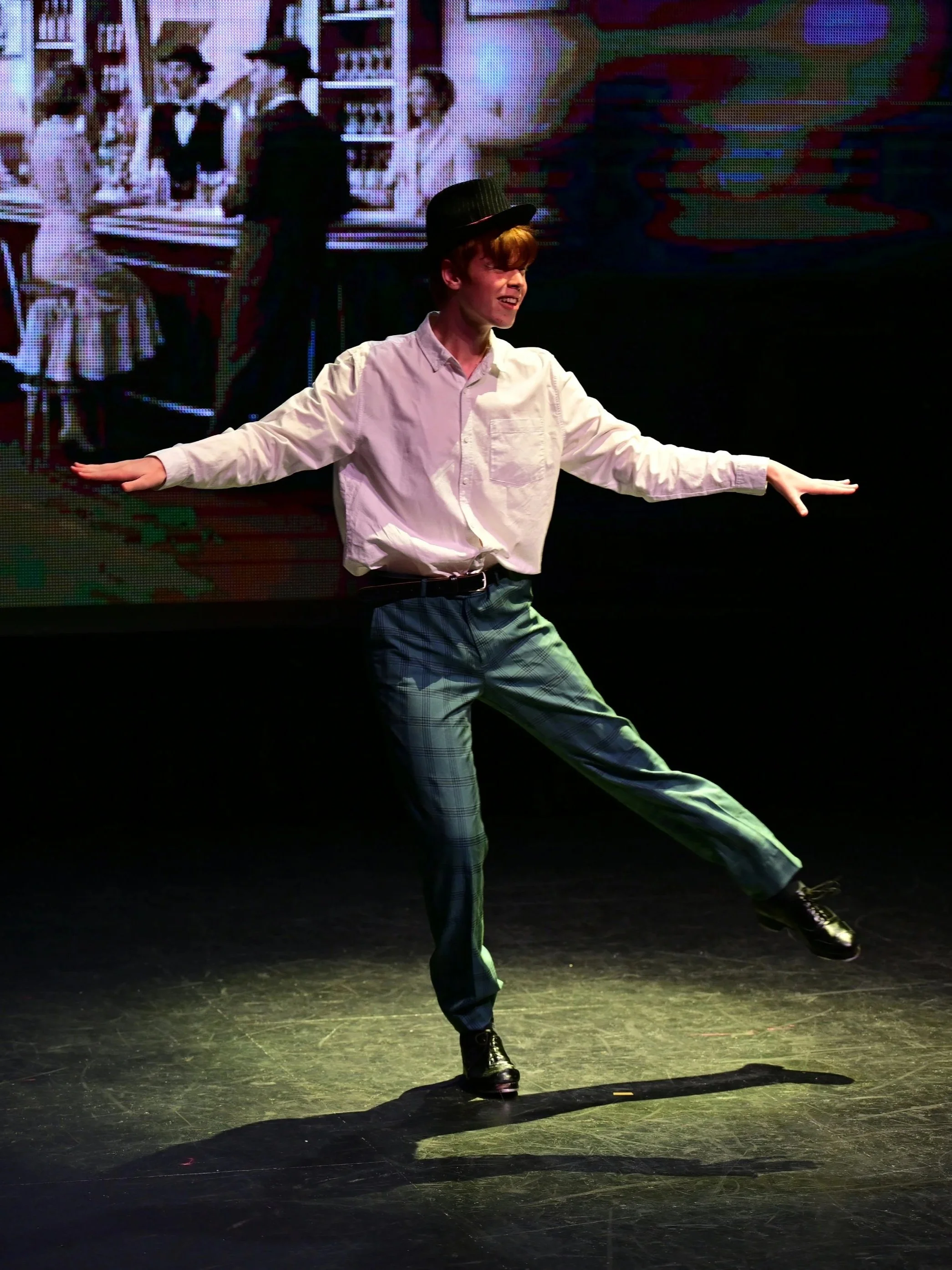 A performer dancing on stage in costume, wearing a white shirt, plaid pants, black boots, and a black top hat. The background shows a large screen displaying scenes of people in a store or library setting.