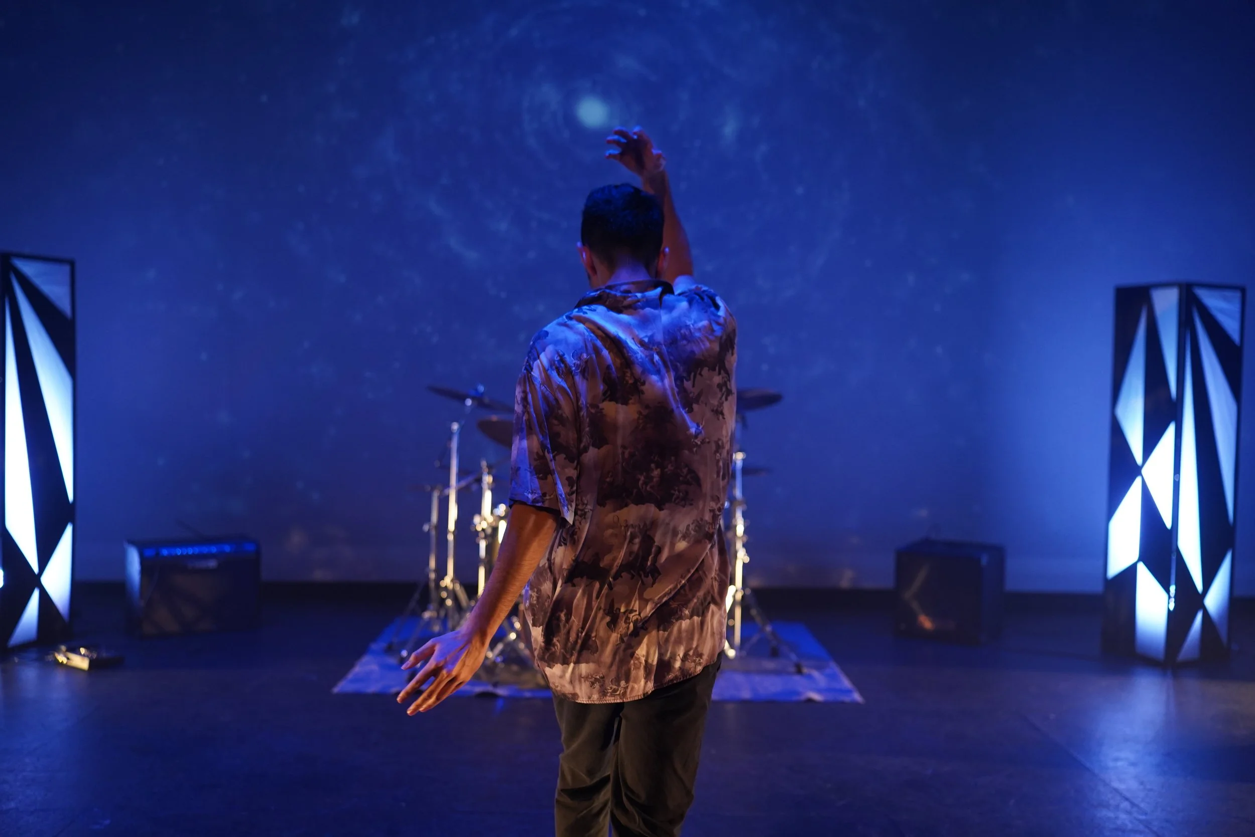 Person dancing or performing on stage with a blue galaxy backdrop, wearing a camouflage shirt, with stage lights and equipment visible.