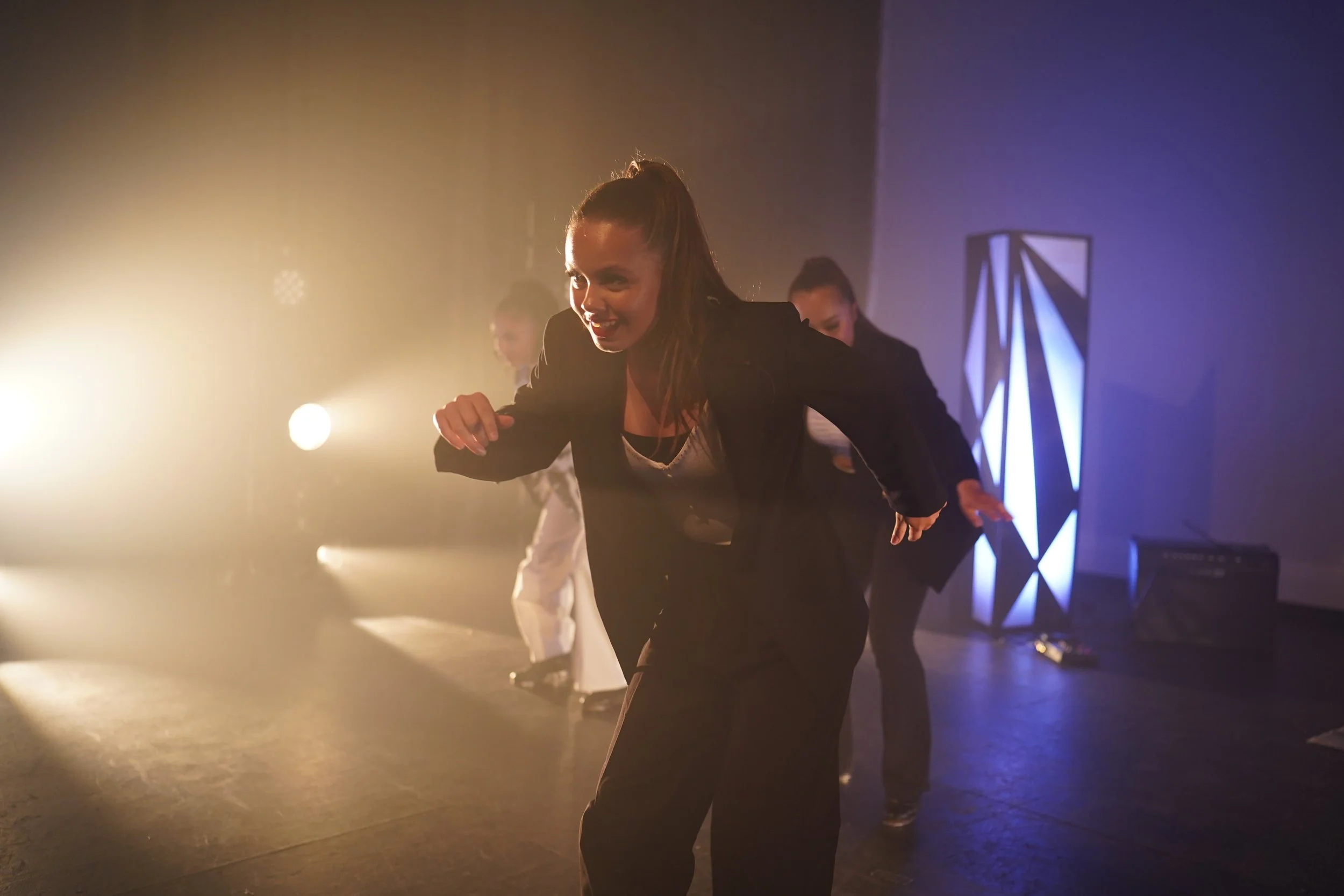 A woman with a high ponytail dancing on stage in a black outfit, illuminated by stage lights, with other dancers in the background.
