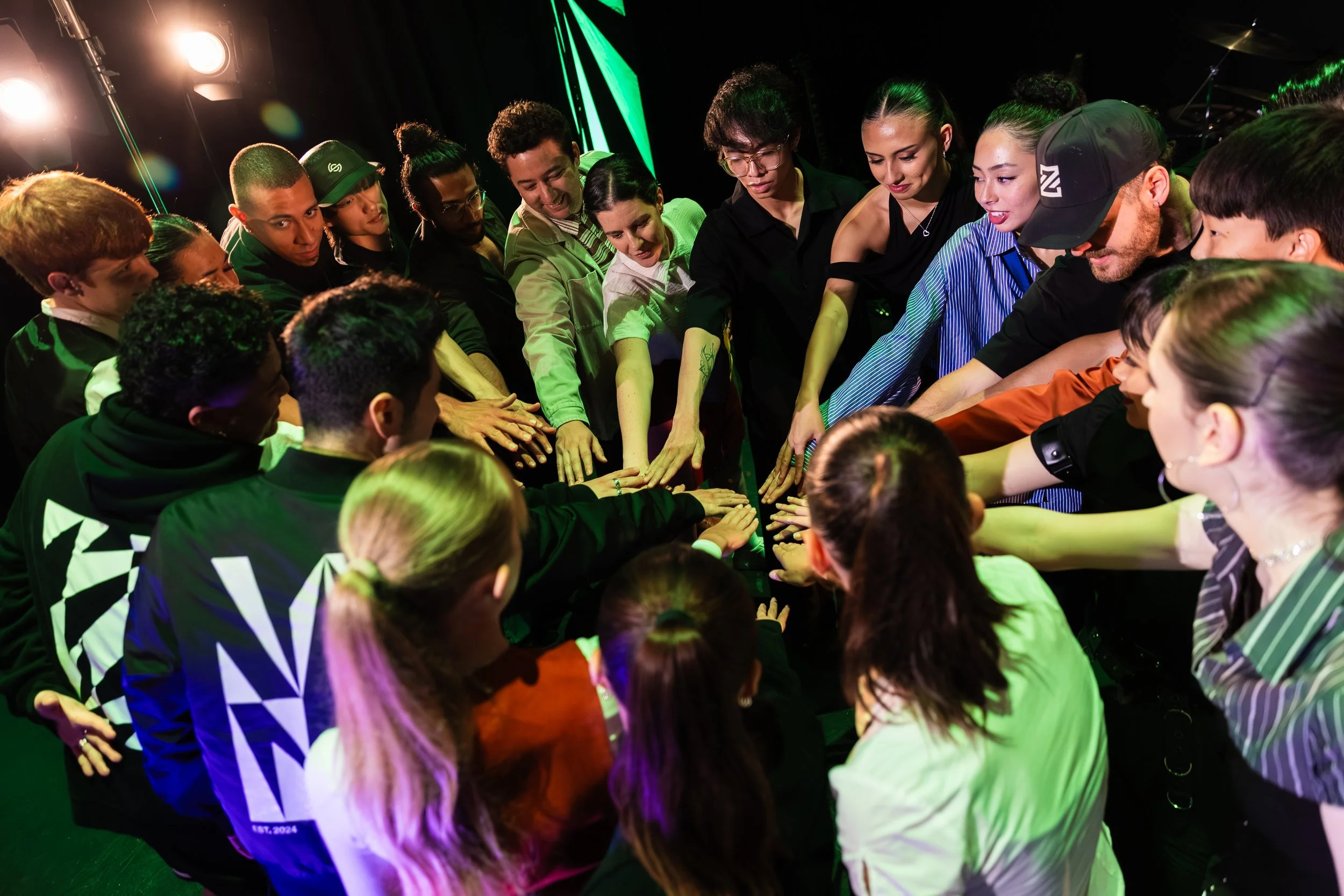 A diverse group of people gathered in a circle with their hands stacked in the center, in a dimly lit room with green and yellow lighting, indicating a collaborative or team-building activity.