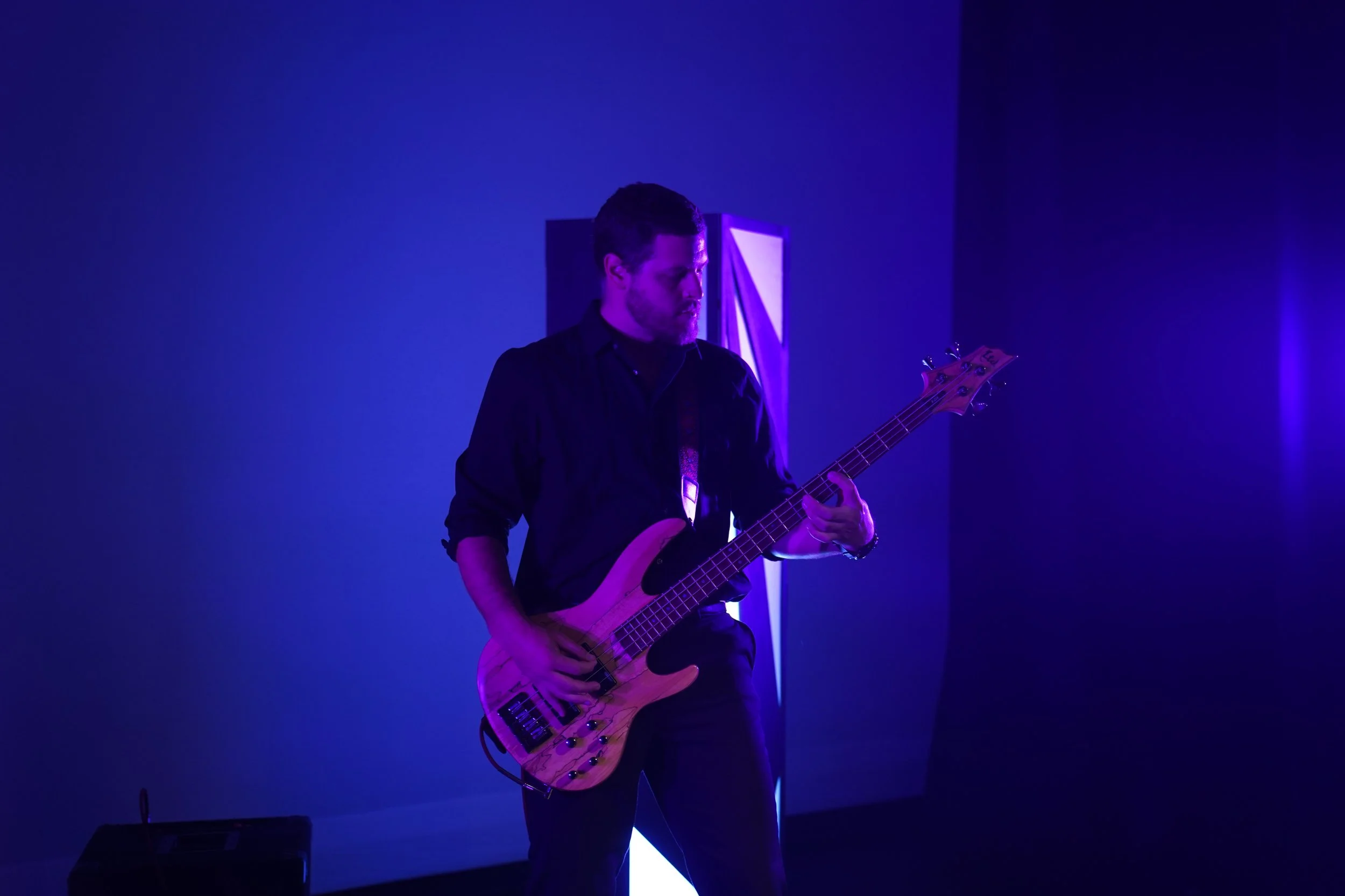 A man playing an electric bass guitar on stage illuminated by purple and blue lights.