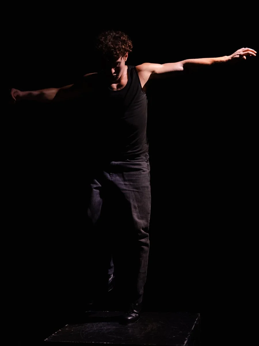 A person standing on a dark platform with arms outstretched, wearing a black sleeveless shirt and dark jeans, against a black background, with dramatic lighting highlighting the figure.