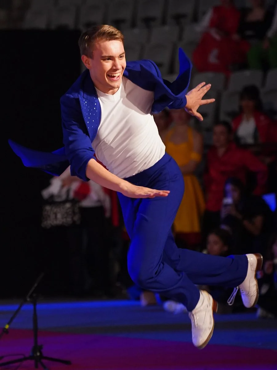 A young man in a white t-shirt and blue suit is jumping mid-air on a stage, smiling, with a microphone stand in the foreground and an audience watching in the background.